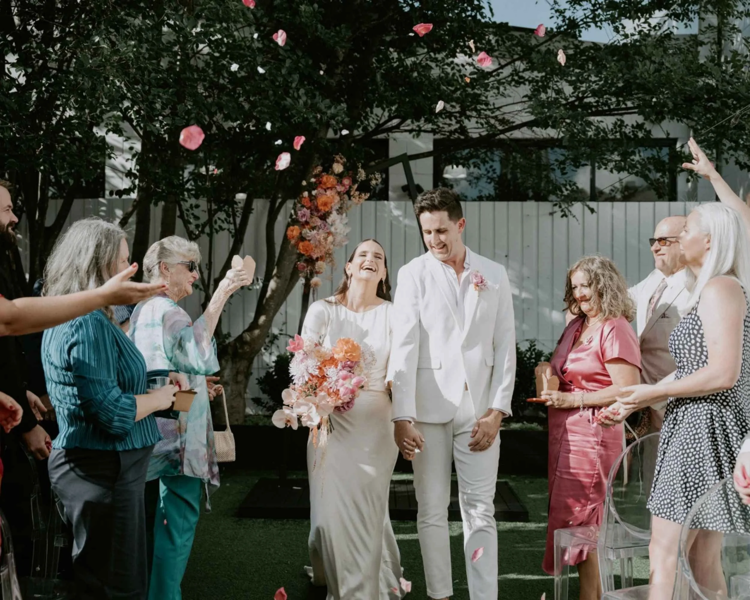 A joyful wedding scene with a bride and groom walking hand-in-hand while guests throw flower petals. The bride is wearing a white gown and holding a bouquet, and the groom is dressed in a white suit. Smiling guests surround them, celebrating in a gar