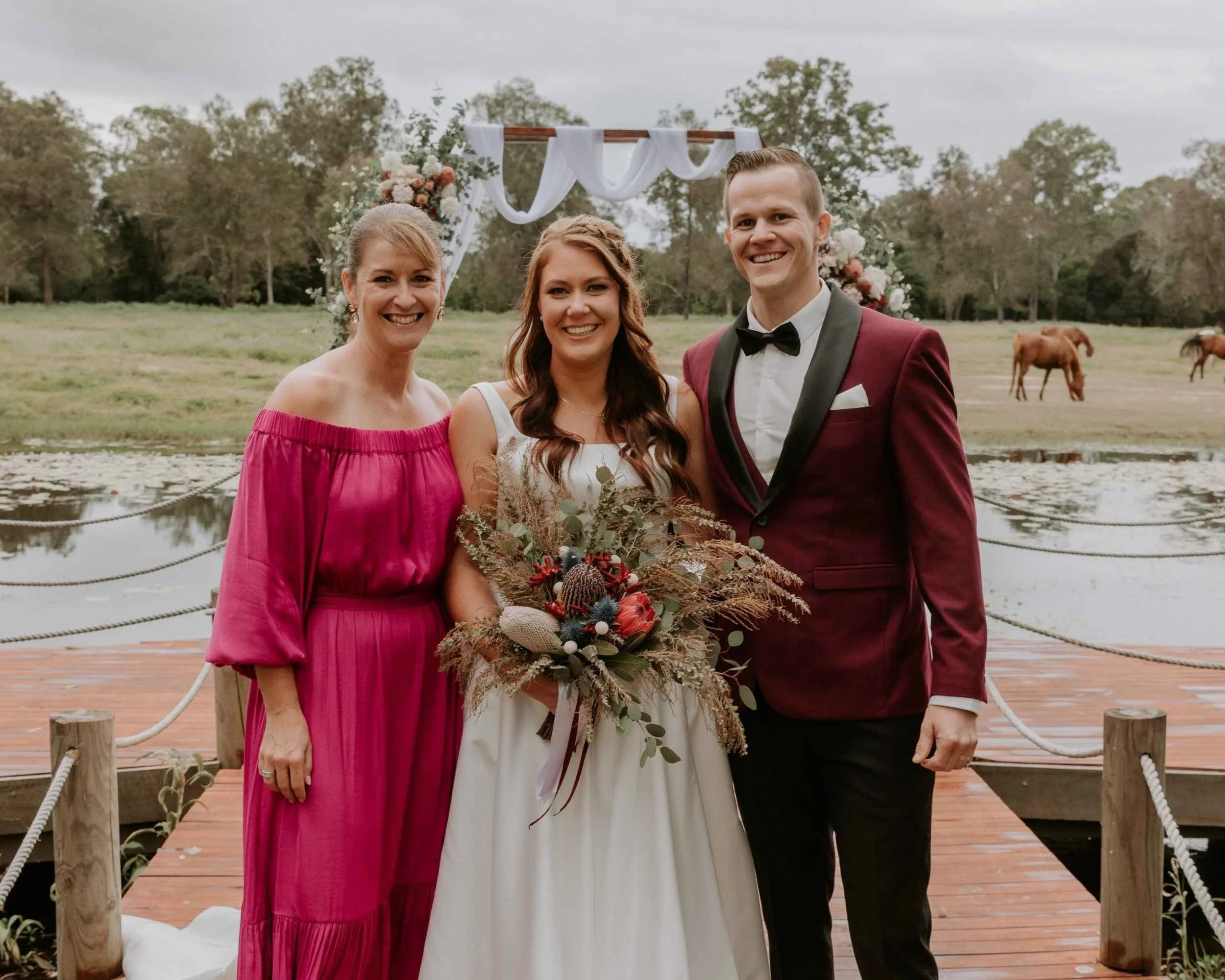 Group portrait at a wedding, featuring two women in wedding attire and one man in a suit, standing on a wooden deck by a pond with horses grazing in the background.