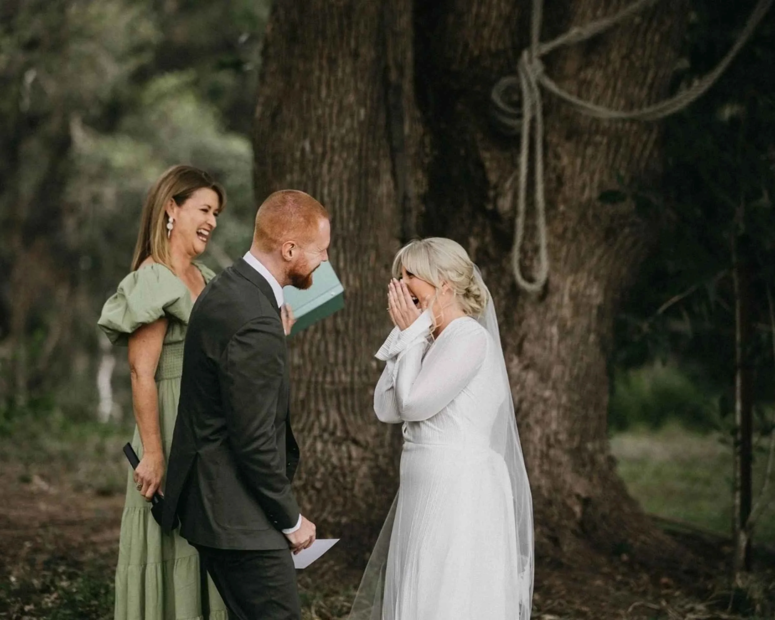 A wedding ceremony in a forest setting with a bride in a white dress, a groom in a black suit, and an officiant in a green dress, all laughing in front of a large tree.