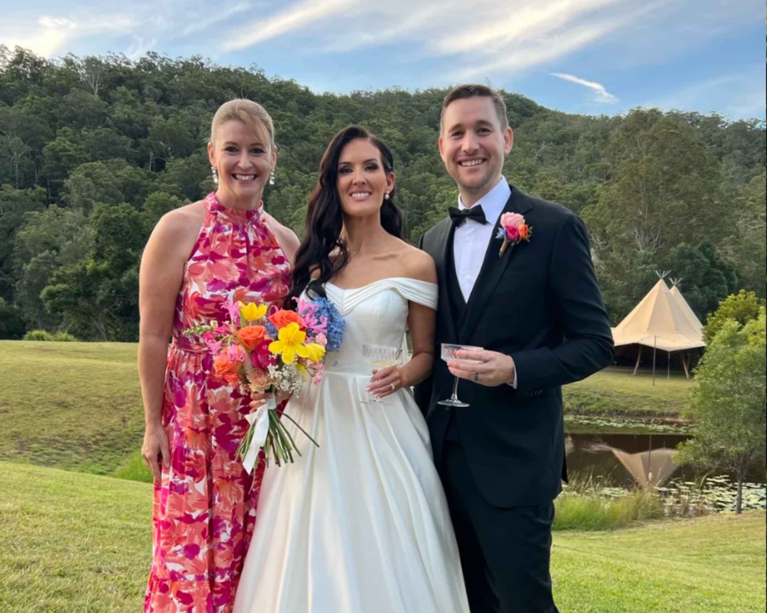 A bride in a white dress holding a bouquet stands between a woman in a pink floral dress and a man in a black suit with a bow tie, all smiling and standing on grass with a scenic outdoor background.