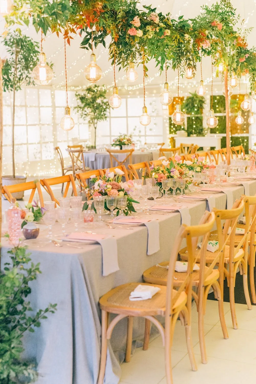 Elegant dining table with wooden chairs, floral arrangements, hanging greenery with string lights, and decorative light bulbs, set in a well-lit room.