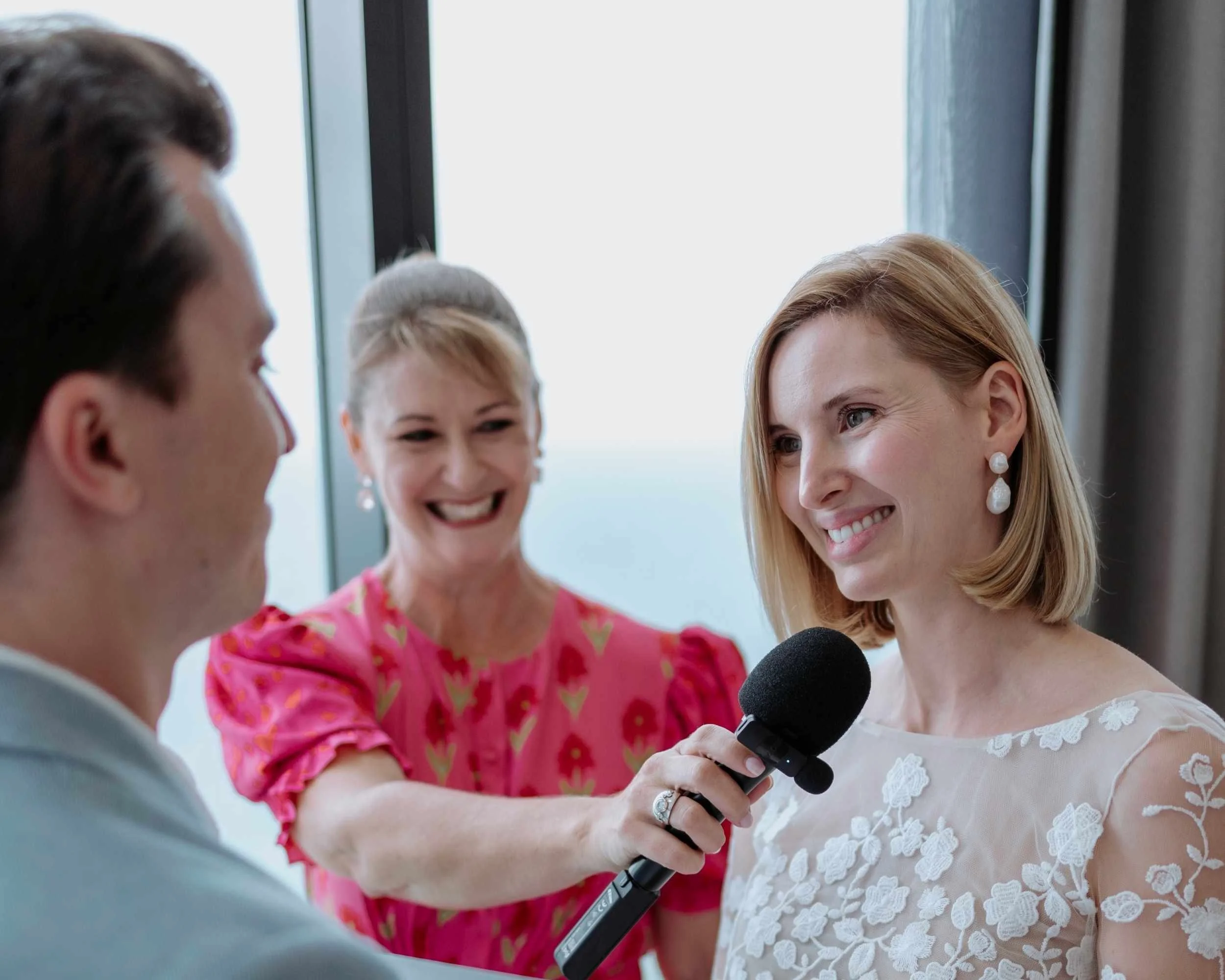 Bride in lace dress holding a microphone and paper, smiling at groom during the wedding ceremony, with officiant in pink dress in the background.