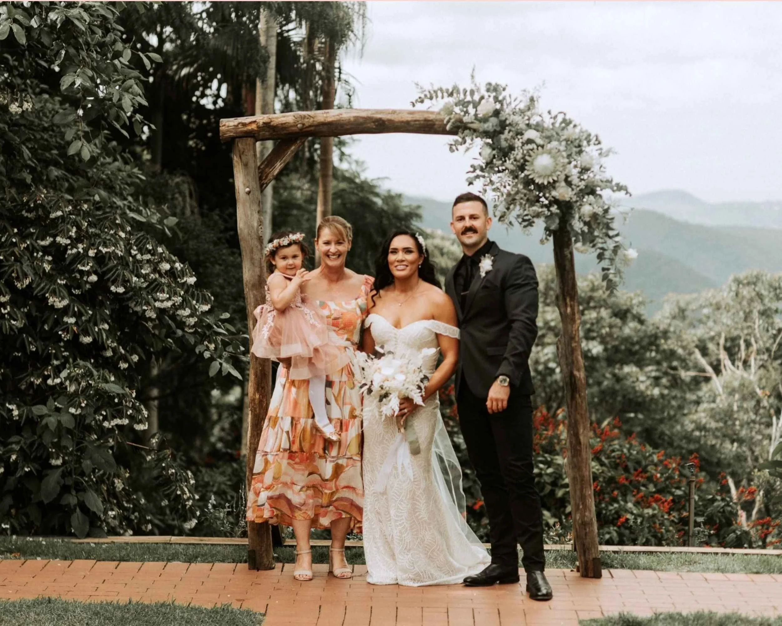 Wedding party posing outdoors under rustic wooden arch with bride, groom, and family members; lush greenery and mountains in the background.