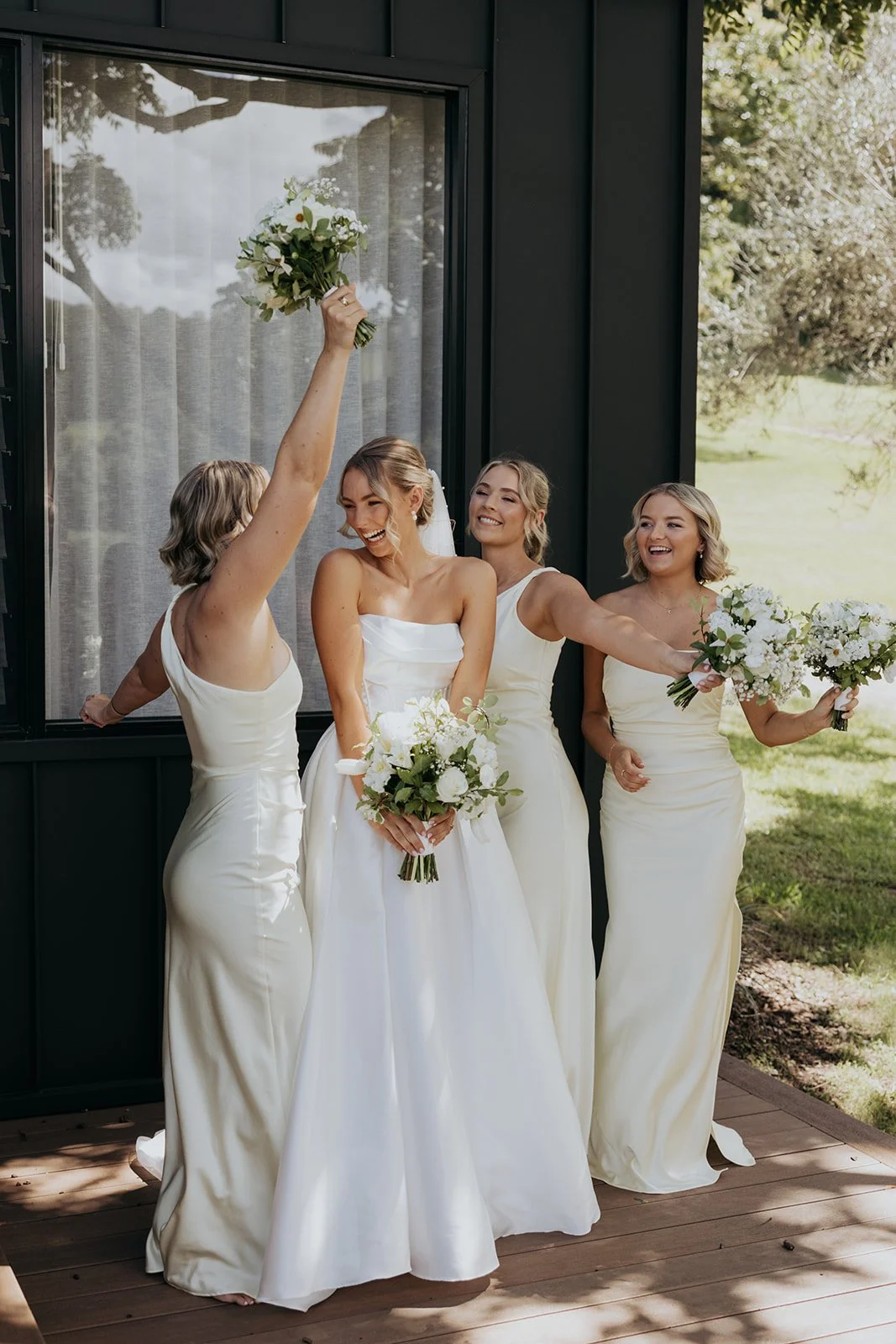 Bride in a white wedding gown with three bridesmaids in cream-colored dresses, celebrating outdoors with bouquets of white flowers, near a dark building with a window, in a lush green setting.