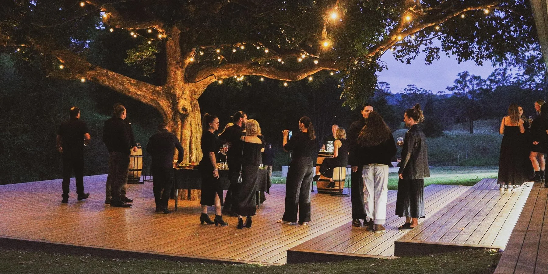 An outdoor evening gathering with people socializing on a wooden deck under a large illuminated tree, with string lights hanging in the branches.