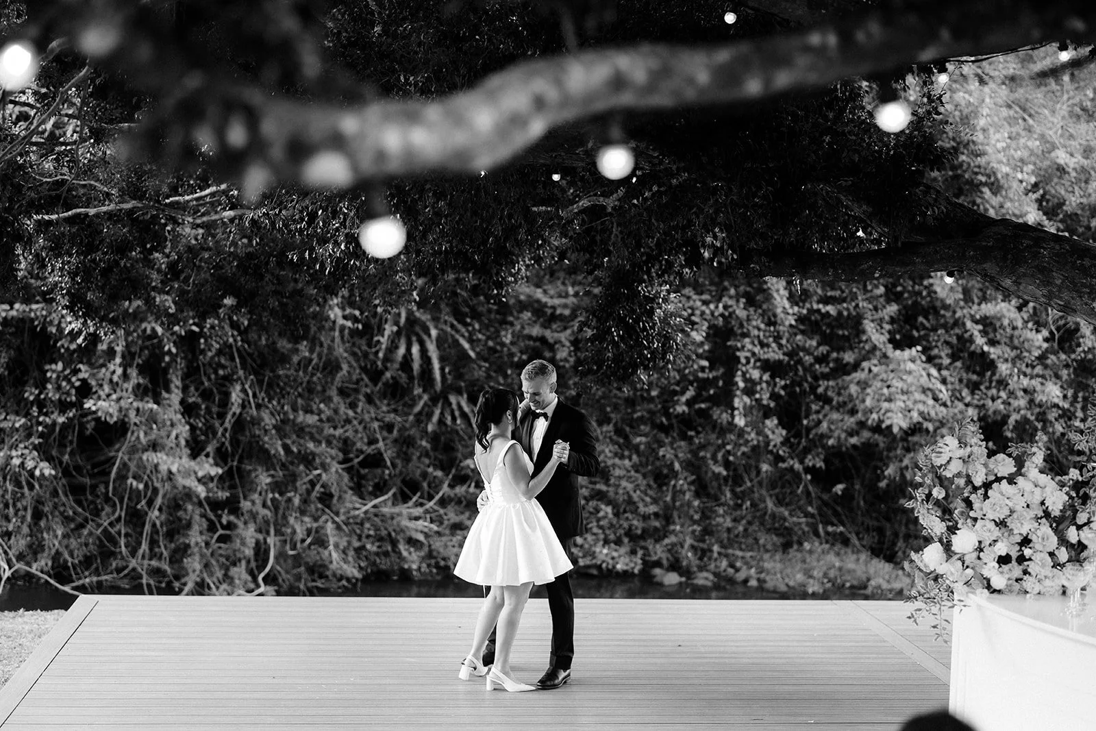 A black and white photo of a couple dancing on an outdoor wooden platform, surrounded by trees and string lights, during their wedding.