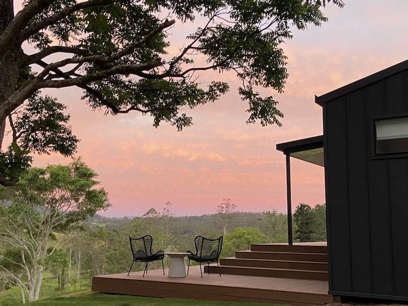 A deck with two outdoor chairs and a small table, set against a lush green landscape and a sunset sky with pink and orange clouds.