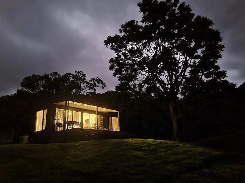 A modern house with large glass windows illuminated from inside, situated on a grassy hill at dusk, with tall trees and a cloudy sky in the background.