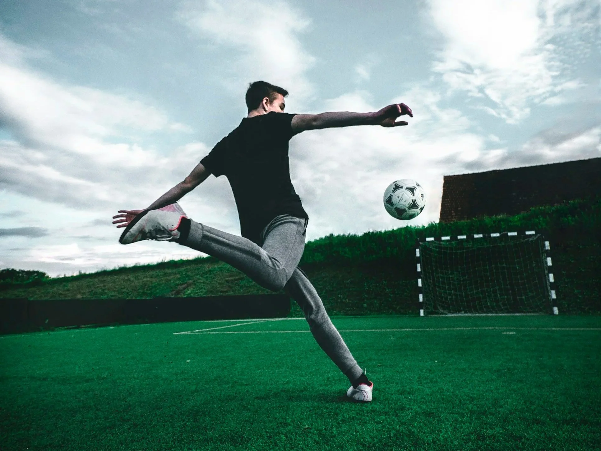 A person in athletic clothing is kicking a soccer ball on a green field with a goal in the background, under a partly cloudy sky.