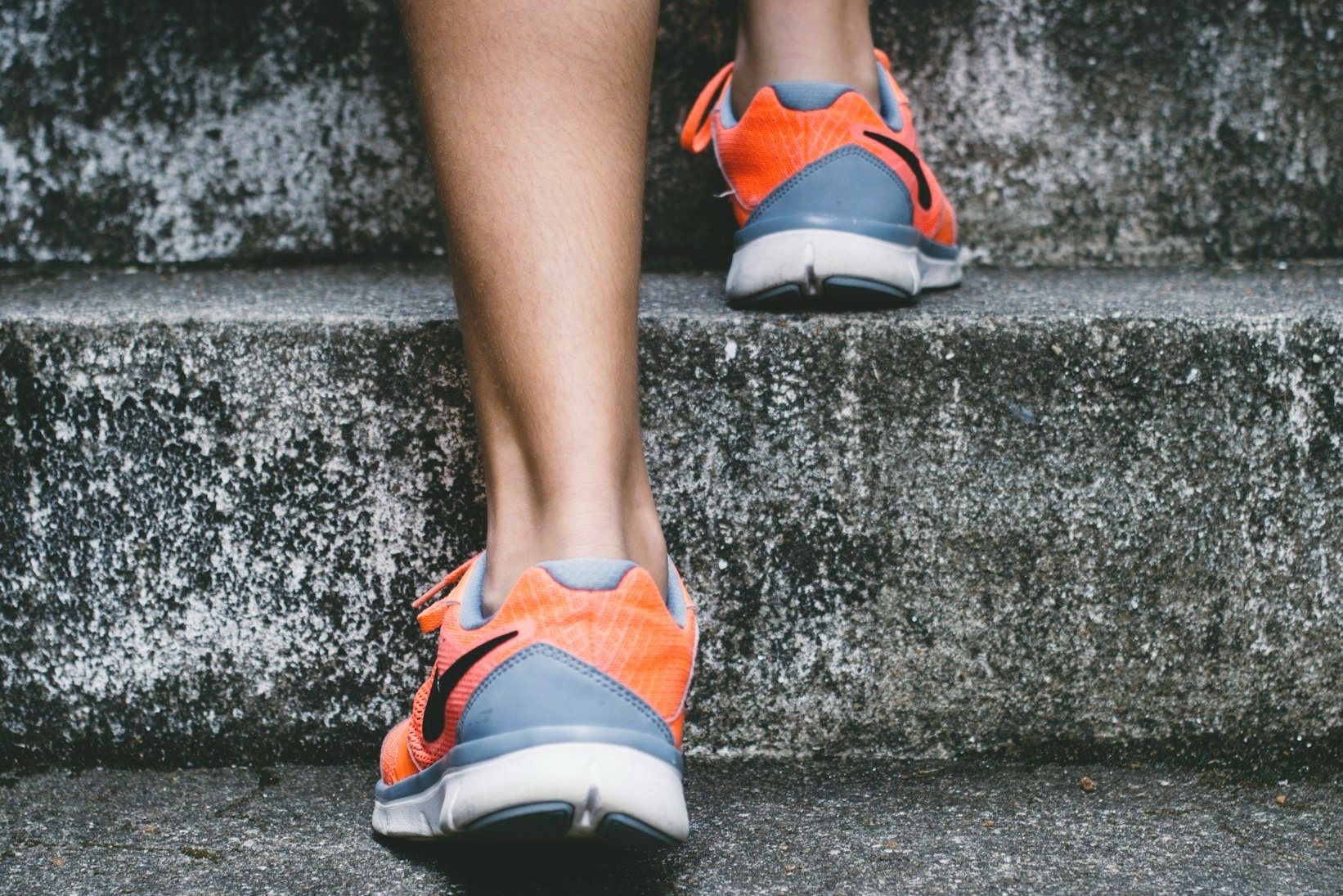 Close-up of a person's legs and feet as they walk up concrete stairs, wearing bright orange and gray running shoes.