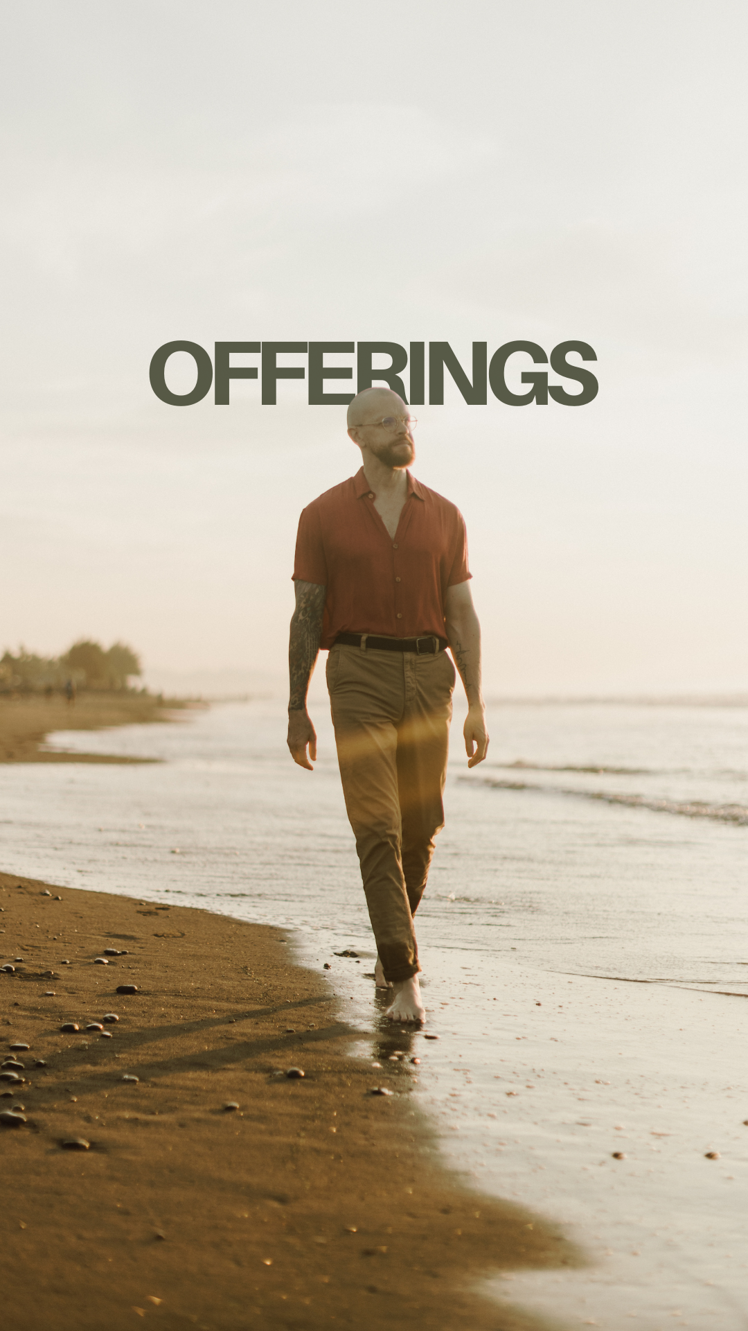 A man with tattoos walking on the beach at sunset, wearing a red shirt and beige pants.