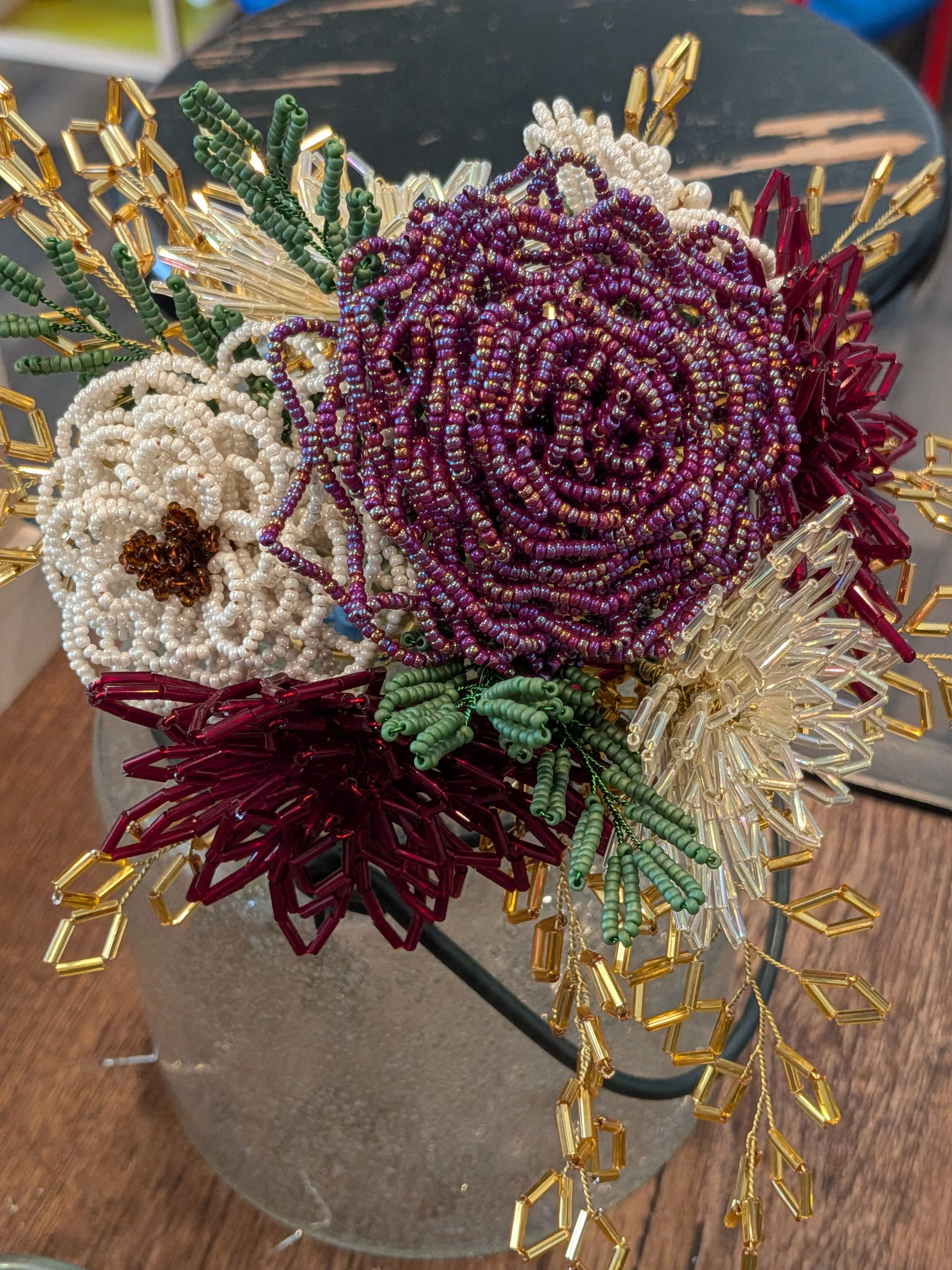 Beaded floral arrangement with purple roses, white flowers, red leaves, green foliage, and gold accents in a glass vase.