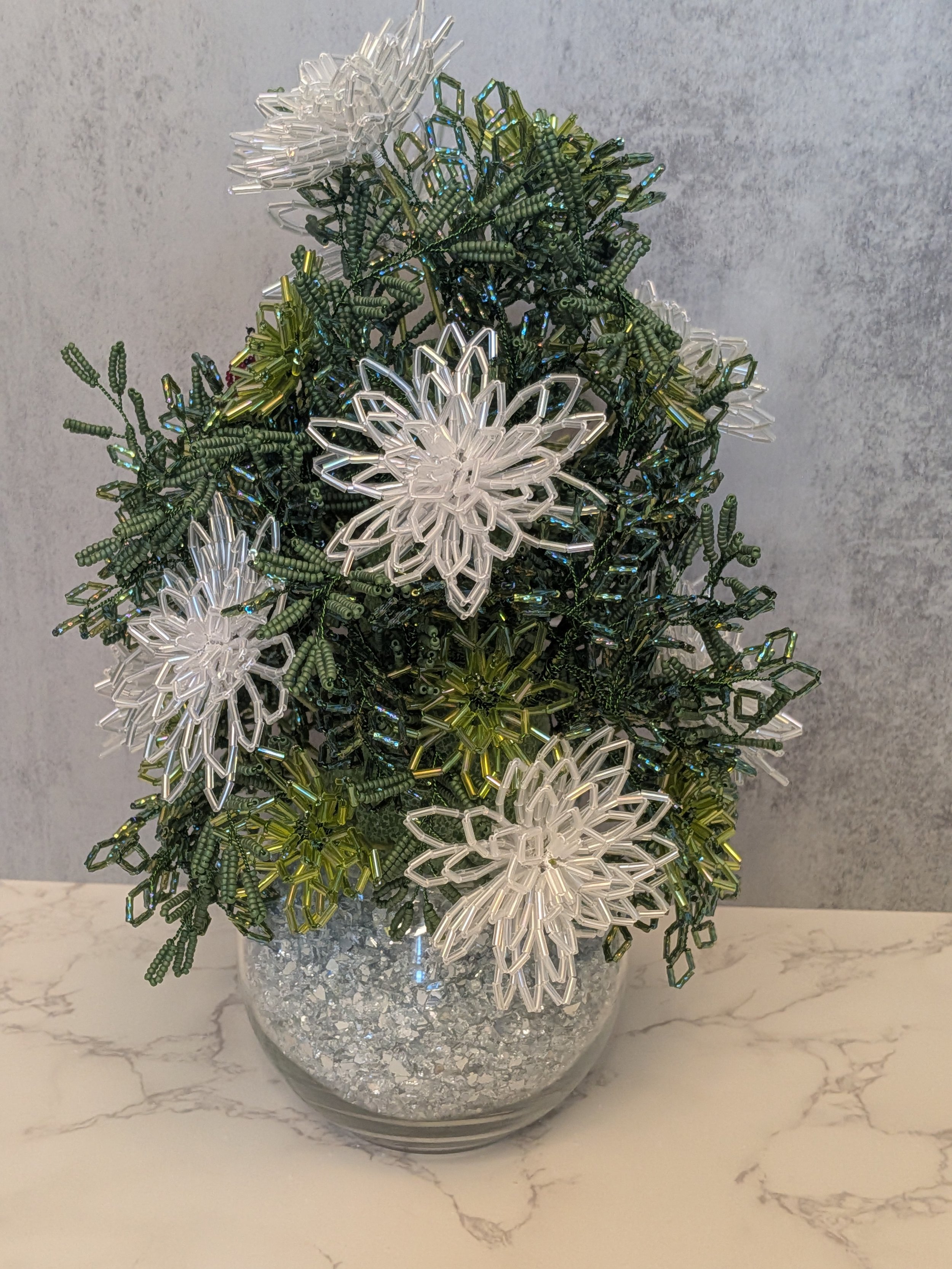 Decorative centerpiece with white and green beaded chrysanthemums in a glass bowl filled with silver glitter, set against a neutral textured background.