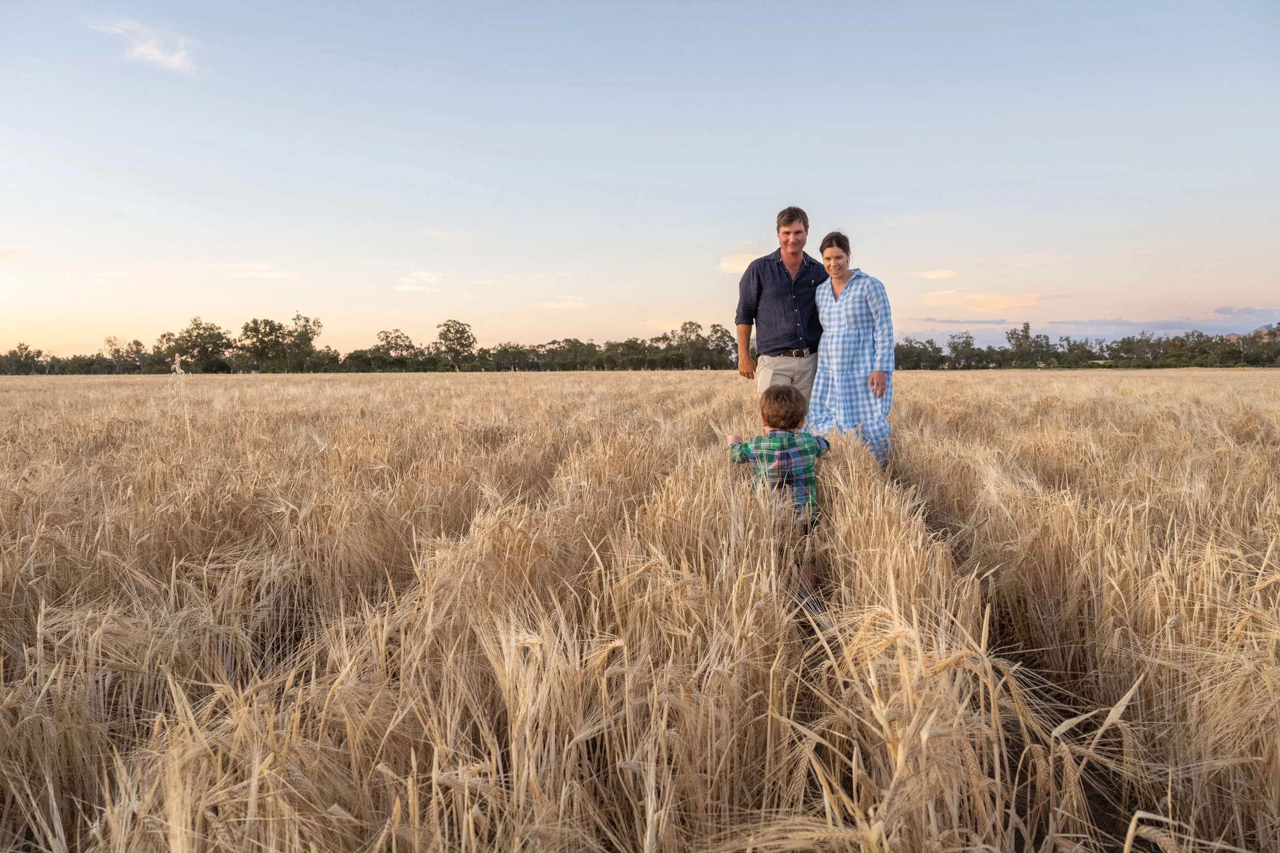 A family of three walking through a golden wheat field at sunset. The man and woman are smiling, and a young child walks ahead of them covering his ears with his hands.