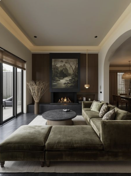 Modern living room with a fireplace, artwork above, green sectional sofa with pillows, round coffee table, large window with curtains, and a view into a dining area.