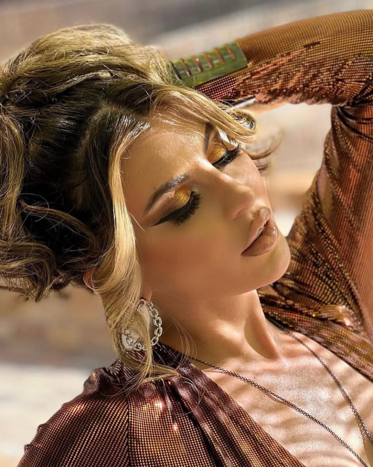 A woman with blonde, wavy hair and dramatic makeup, wearing a shiny copper-colored top and jewelry, posing with her eyes closed.