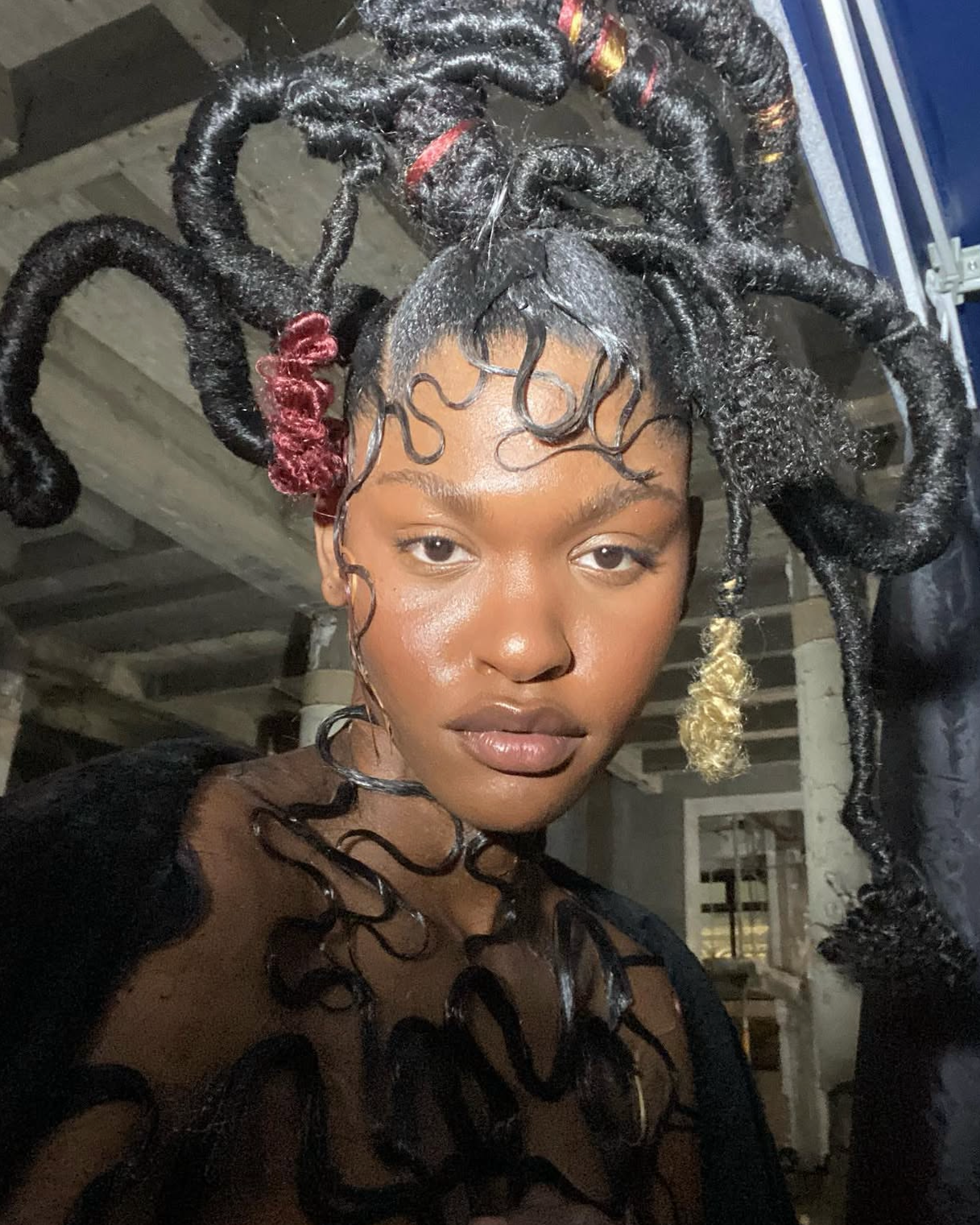 A woman with styled dreadlocks, some with colored tips, wearing a black sheer top with black swirling patterns, standing indoors with a visible ceiling.