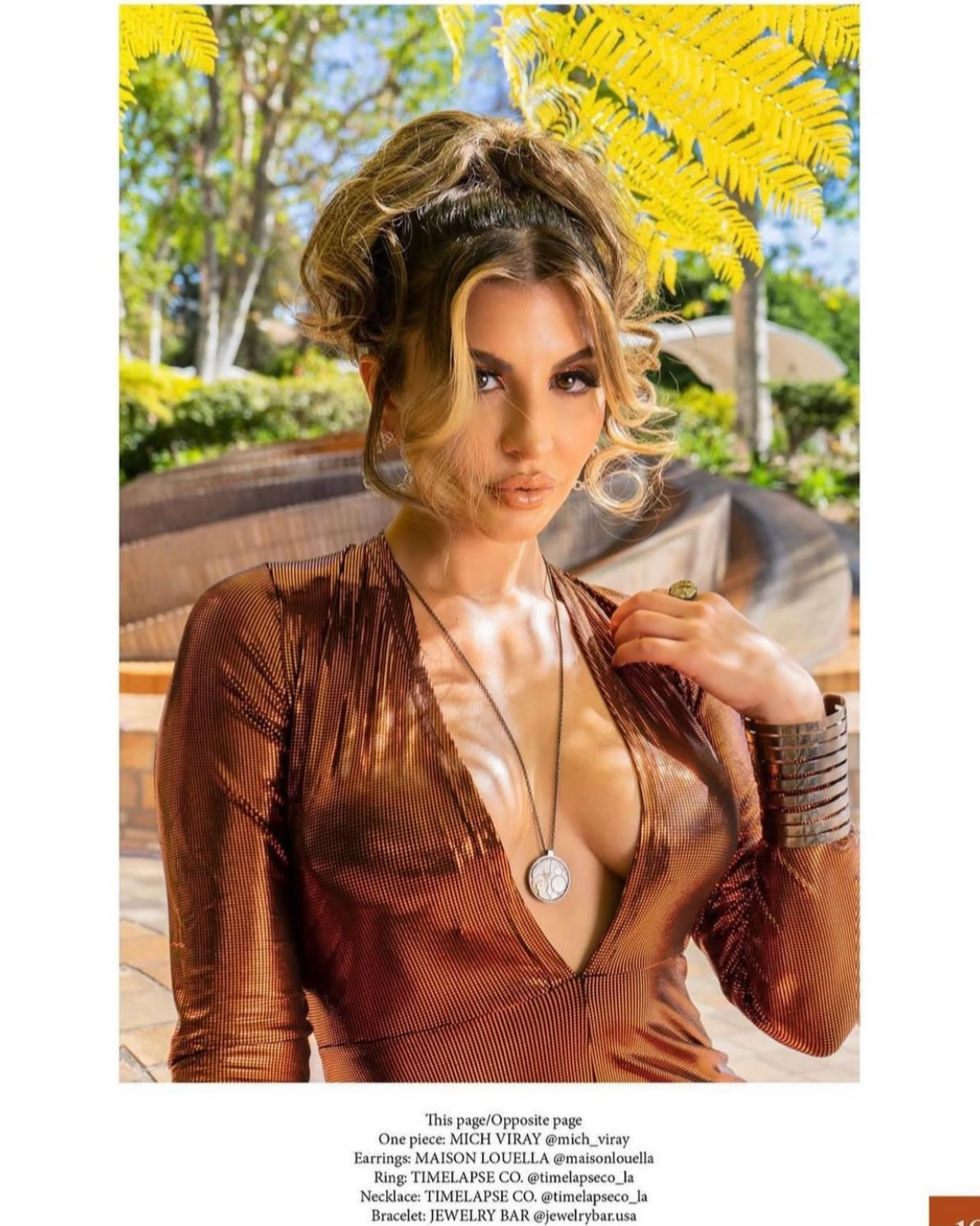 A woman with curly hair wearing a deep V-neck shiny brown top, a round necklace, a large bracelet, and a ring, standing outdoors with lush greenery and yellow leaves in the background.