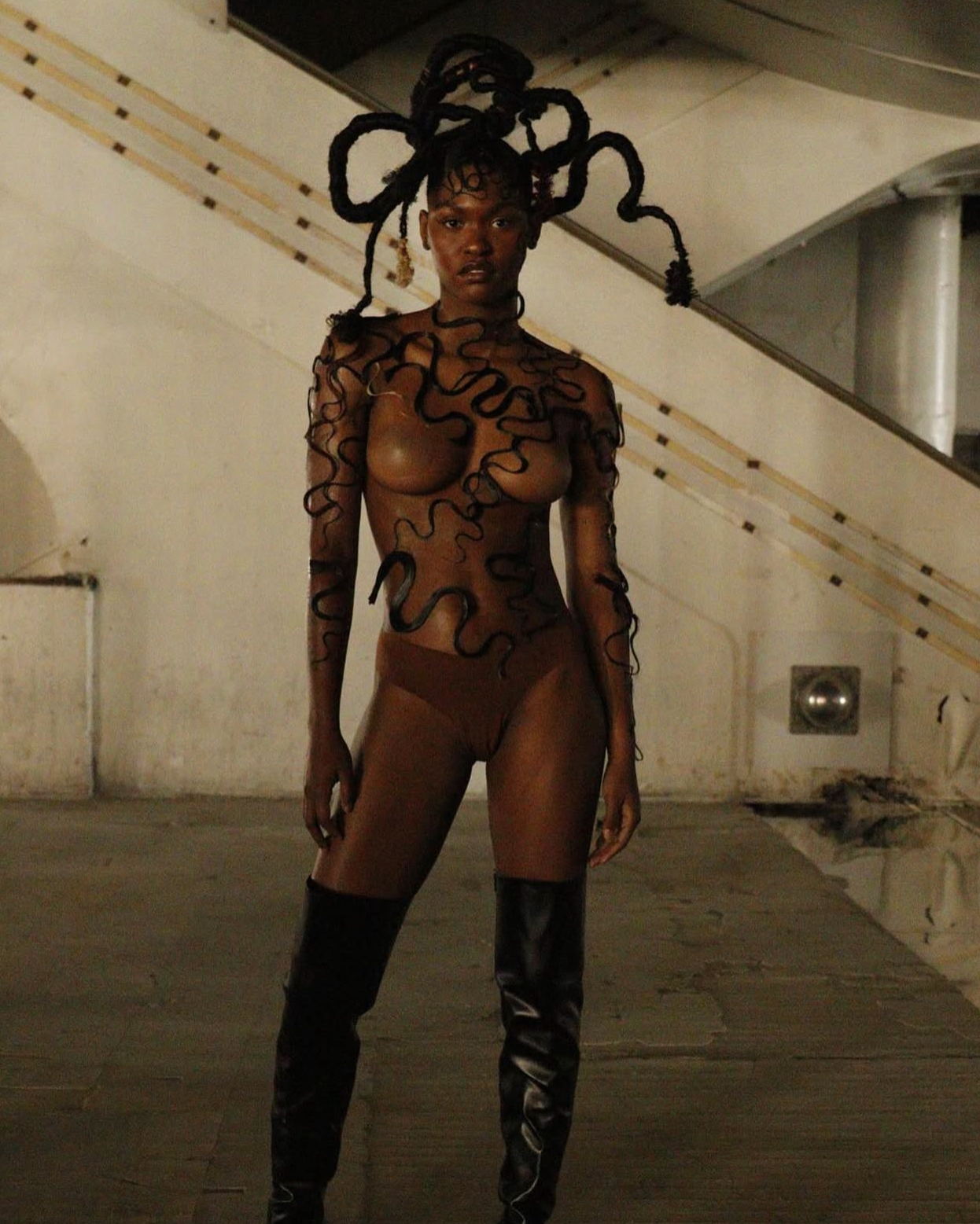 A woman with artistic braided hair, wearing a sheer black outfit with decorative black cords, standing in an industrial-style space with beige walls and a metal staircase.