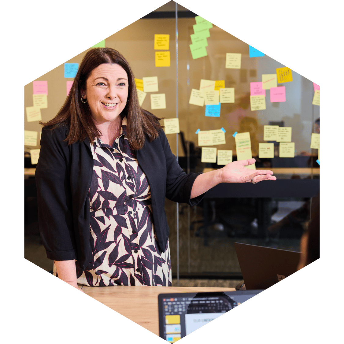 Woman smiling and gesturing in front of a glass wall covered with colorful sticky notes in a workspace.
