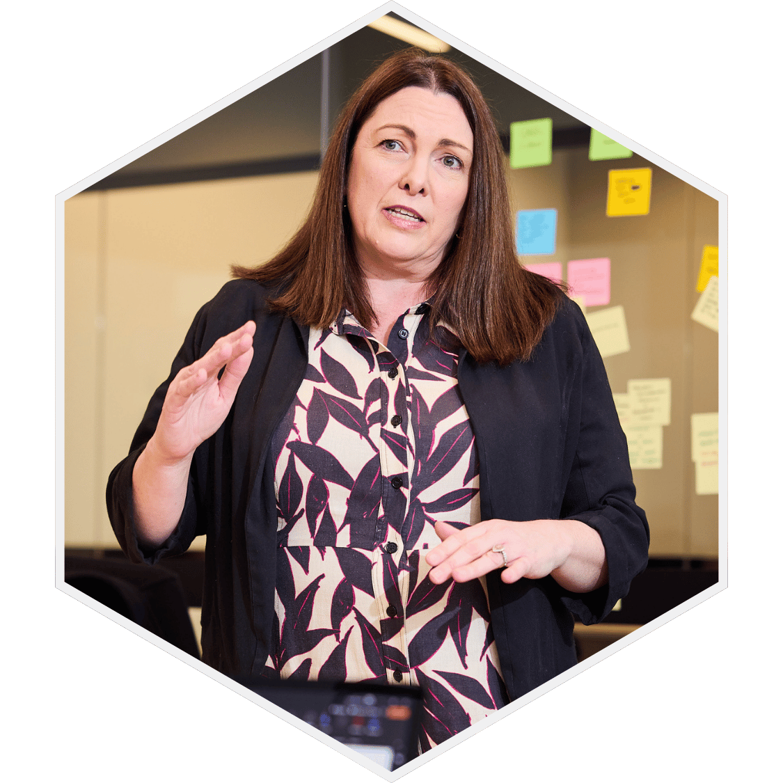 A woman with shoulder-length brown hair, wearing a patterned blouse and black jacket, gestures with her right hand while speaking, in an indoor setting with colorful sticky notes on the wall behind her.