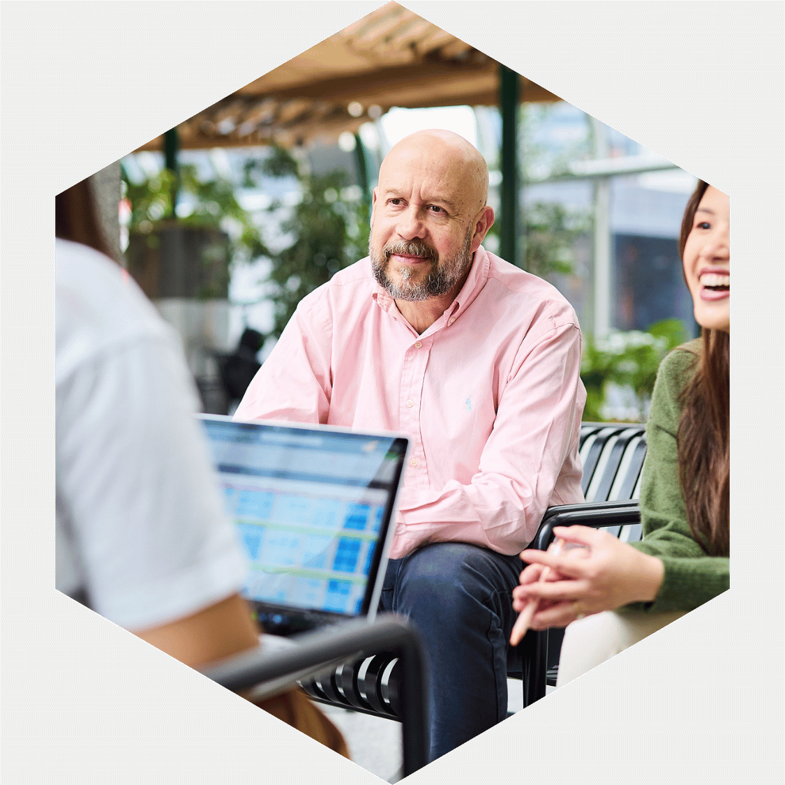 A man with a beard wearing a pink shirt engaged in a conversation with two women, one with long hair in a green sweater and another person with a laptop, in an outdoor setting with greenery.