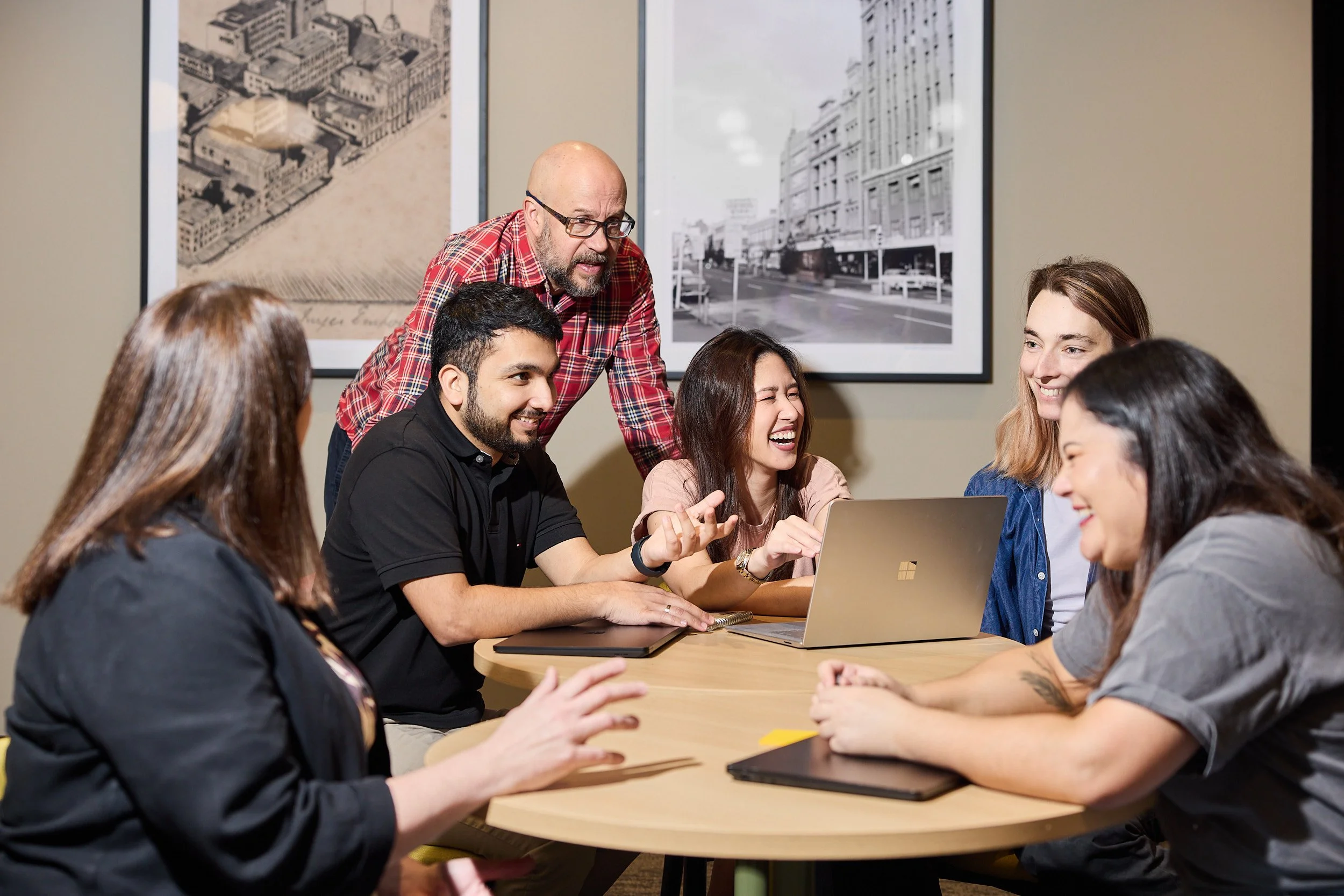 A group of six people sitting around a table with laptops, engaged in a discussion. One man stands, leaning over, speaking to the others who are smiling and laughing. The background features framed black and white photographs of urban scenes.