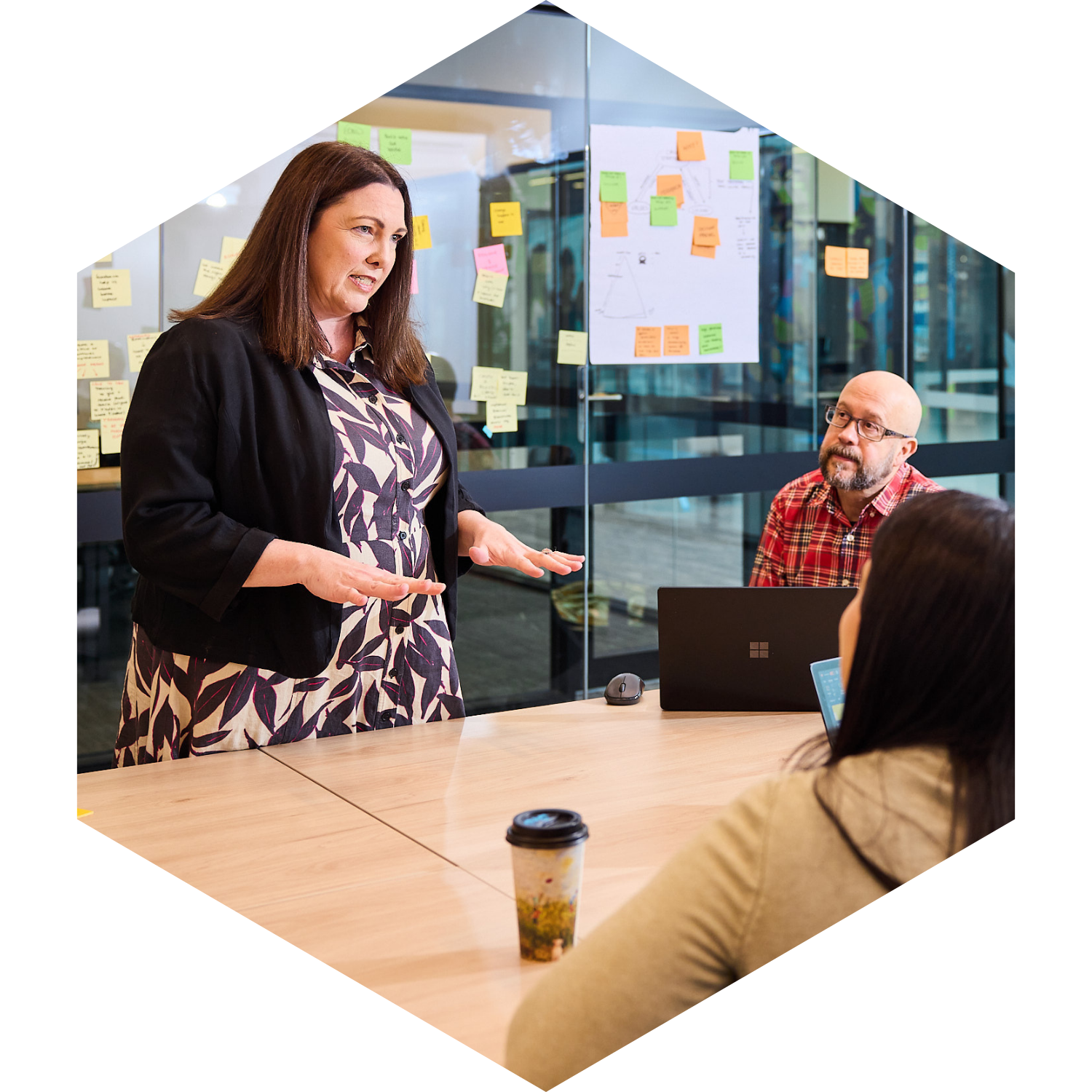 A woman with long brown hair, wearing a black blazer over a patterned dress, is speaking to three other people in a modern office conference room. One person, a man with glasses and a beard in a red plaid shirt, is seated at the table, while another woman with dark hair is partially visible, sitting across from him. Post-it notes and a large sheet of paper with diagrams are on the glass wall behind them. A takeout coffee cup is on the table.