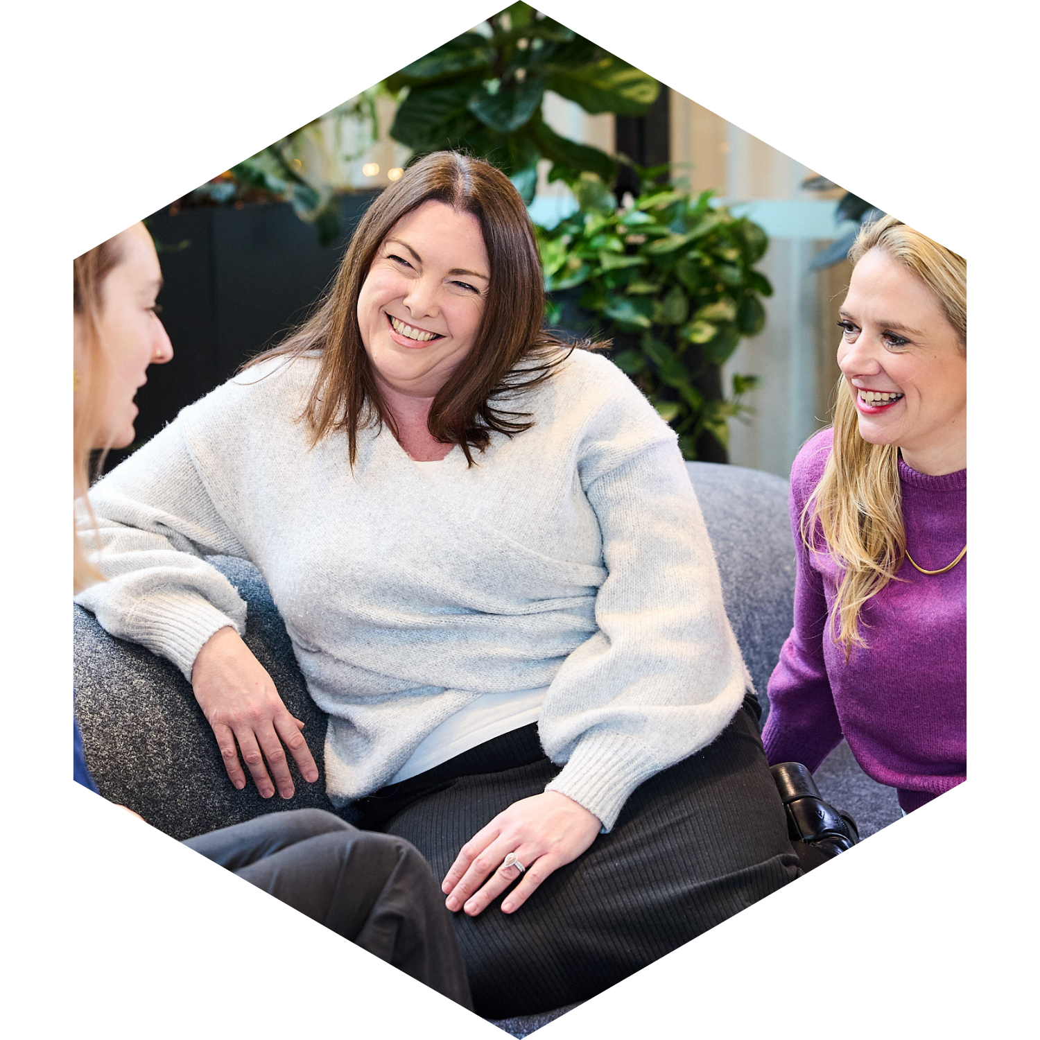 Three women sitting on a gray couch and engaging in conversation, smiling and laughing, indoors with plants in the background.