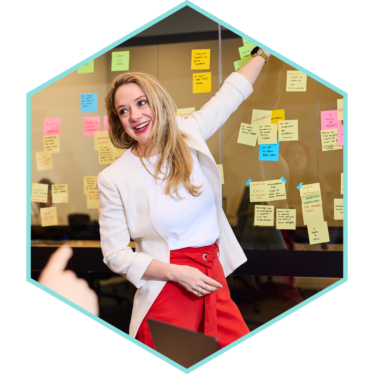 A woman with long blonde hair wearing a white blazer and red pants standing in front of a glass wall covered with colorful sticky notes, smiling and pointing at the notes.