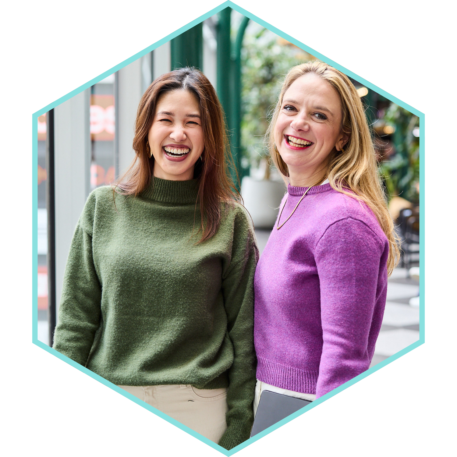 Two women smiling, one with long brown hair wearing a green sweater, and the other with blonde hair wearing a purple sweater, standing indoors with a blurred background of greenery and furniture.