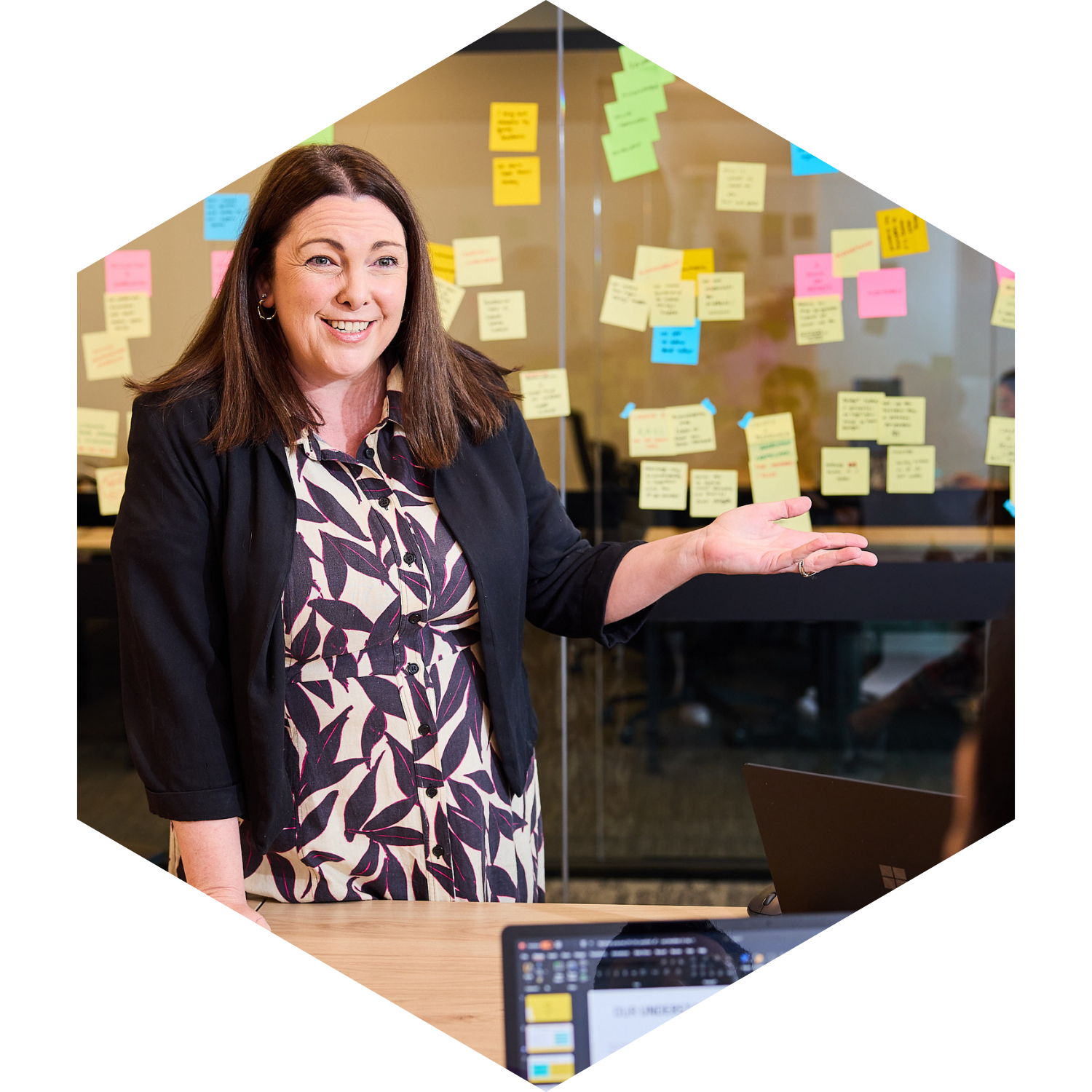A woman standing in front of a glass wall covered with colorful sticky notes, explaining or presenting in an office environment.