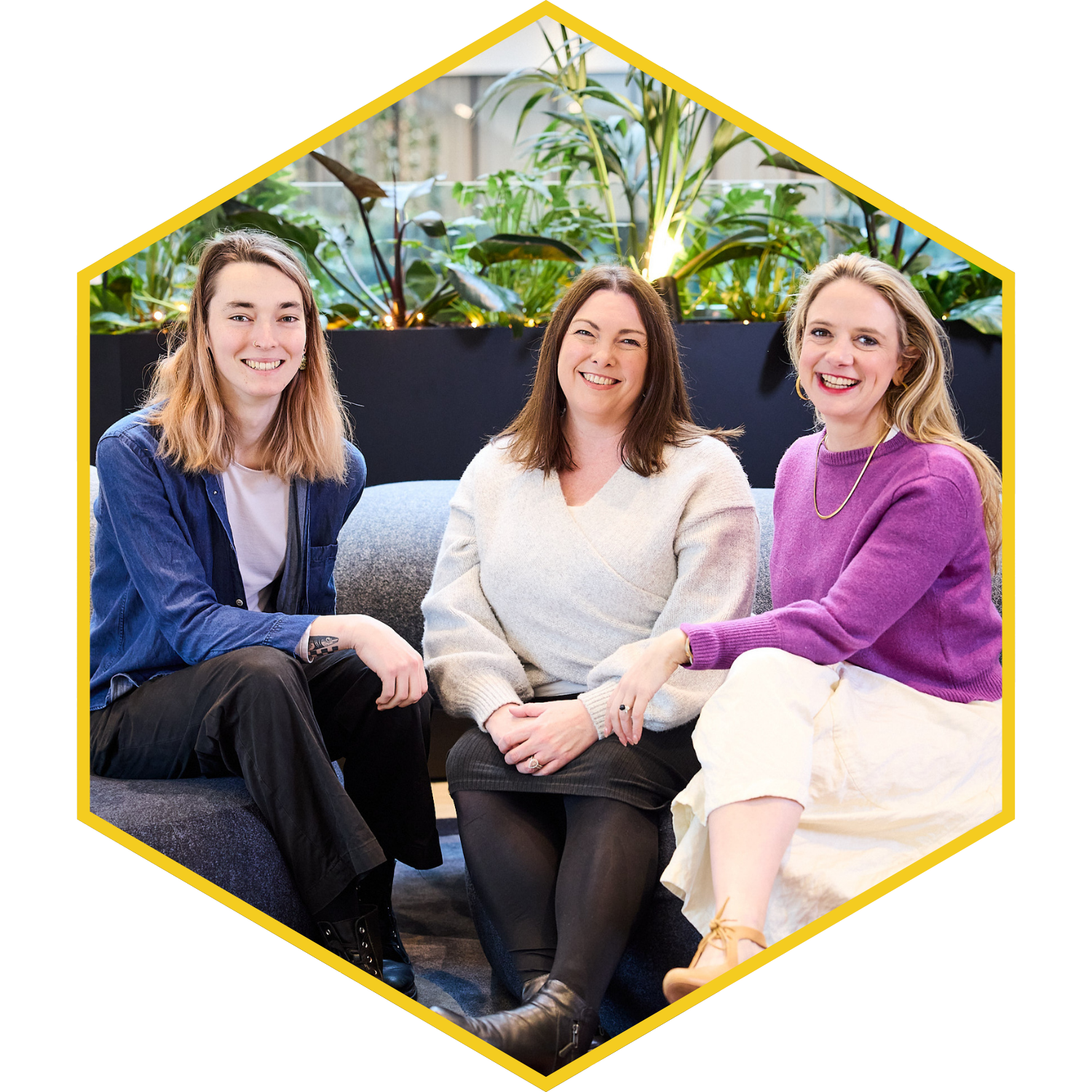 Three women sitting on a couch, smiling, with green plants in the background.