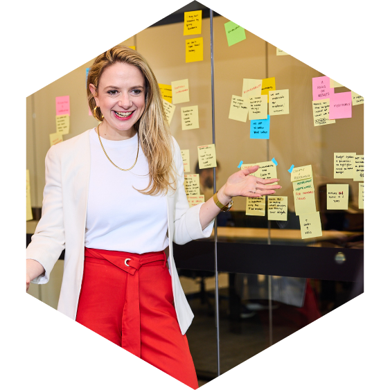 A woman in a white blazer and red pants smiling and gesturing in front of a glass wall covered with colorful sticky notes.