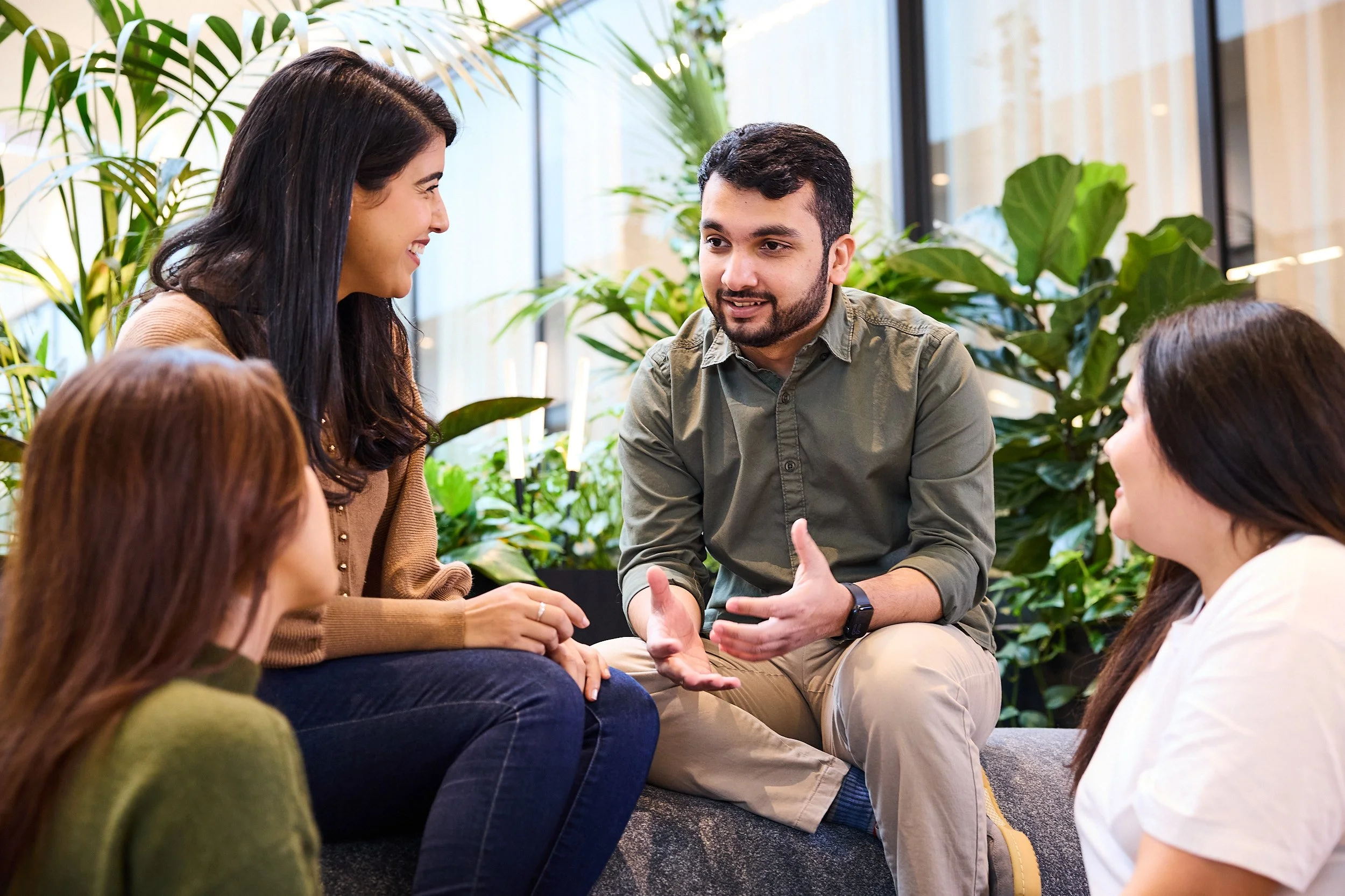 A group of five young adults engaging in conversation indoors, with large green plants and window lighting in the background.