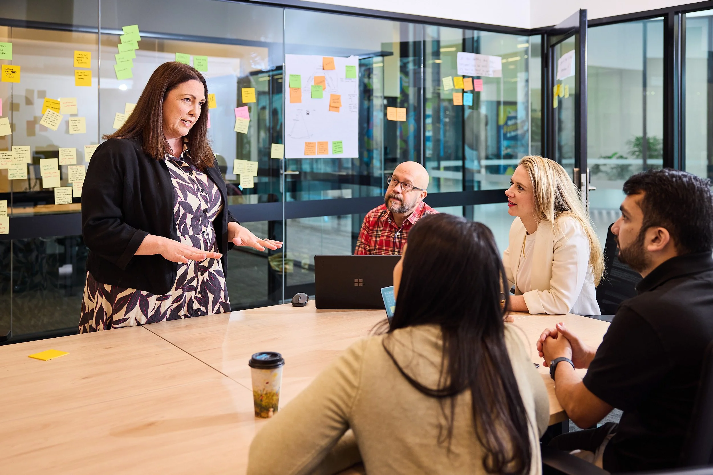 A woman in a black blazer and printed blouse is speaking to a group of diverse people seated around a conference table in a modern office. The group is engaged and attentive. The office has large glass walls with colorful sticky notes and charts posted on them.