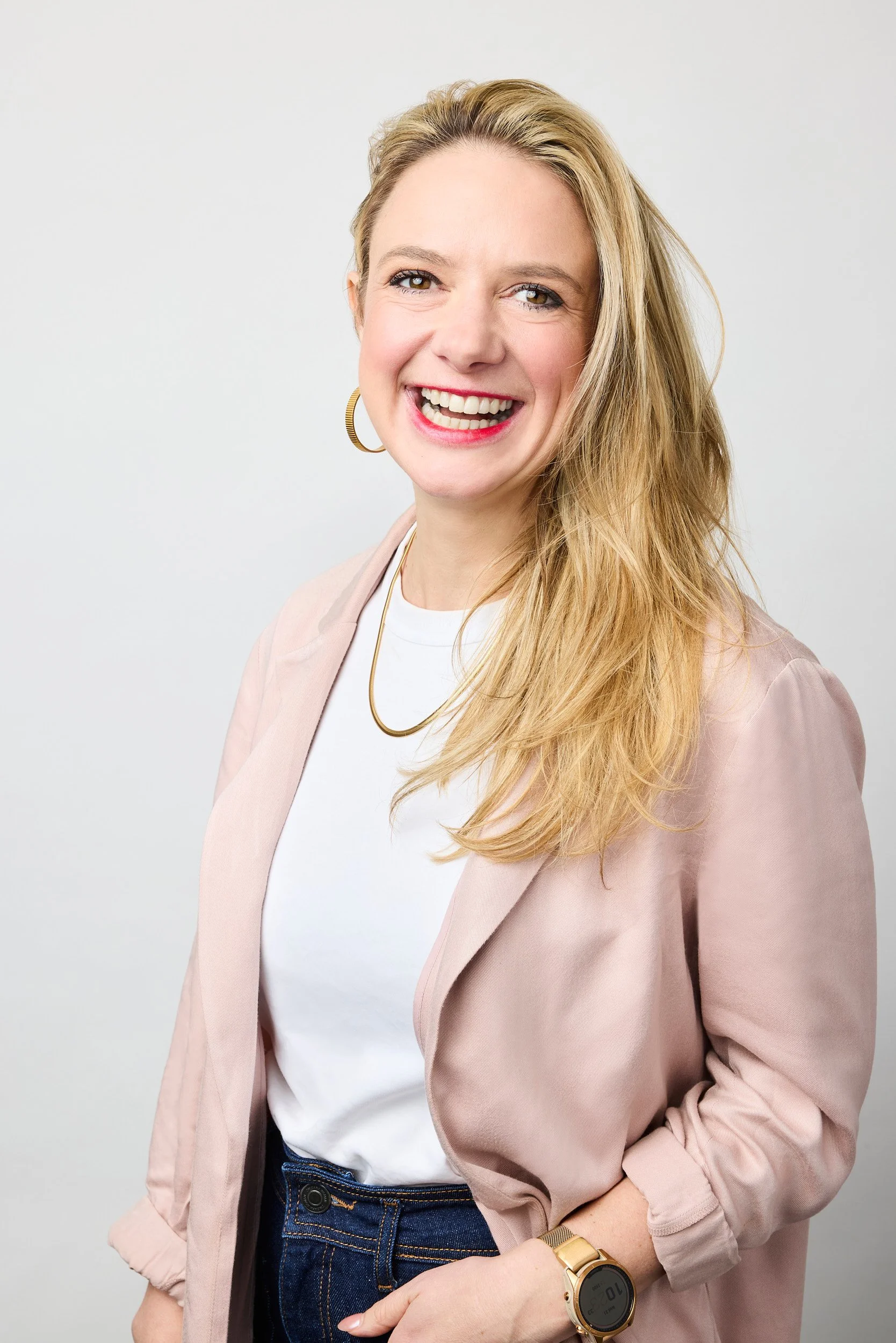 A woman with long blonde hair, wearing a pink blazer over a white t-shirt, with hoop earrings and a gold watch, smiling widely against a plain white background.