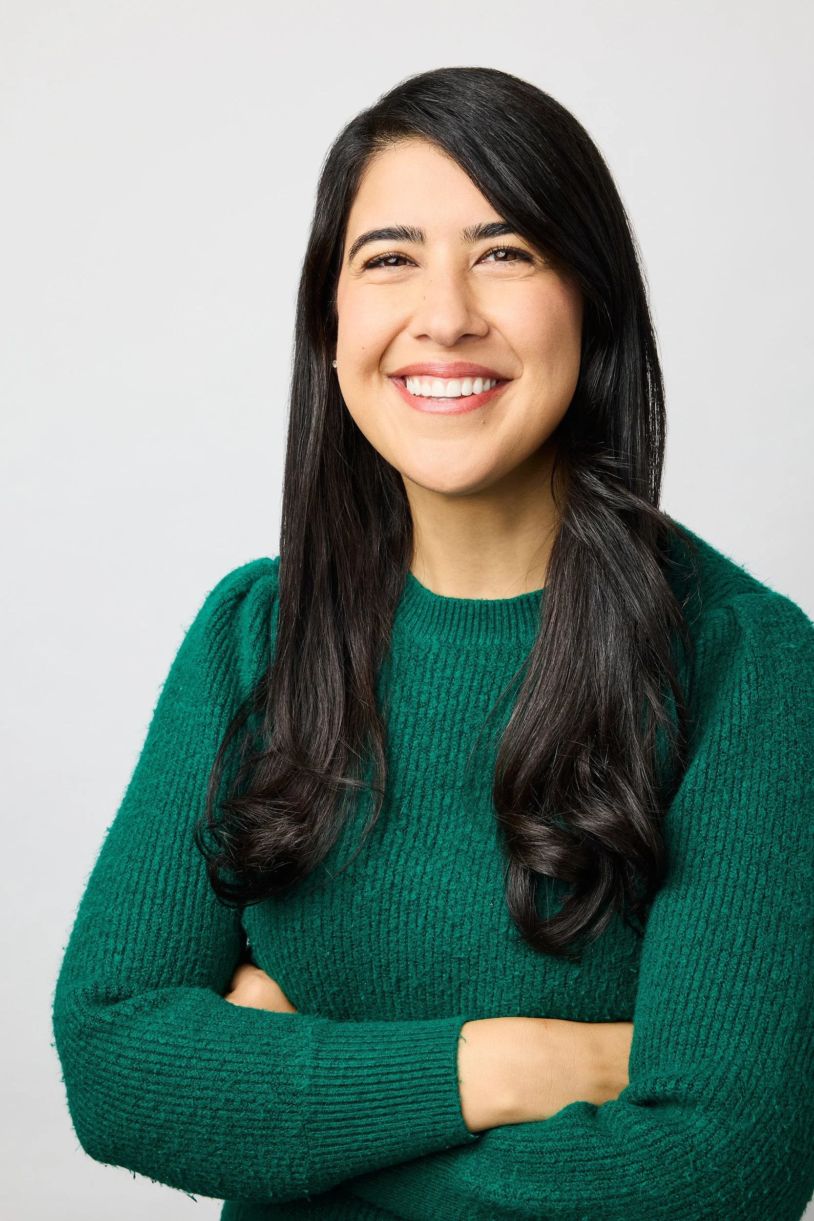 A woman with long dark hair smiling, crossing her arms, wearing a green sweater, against a plain light background.