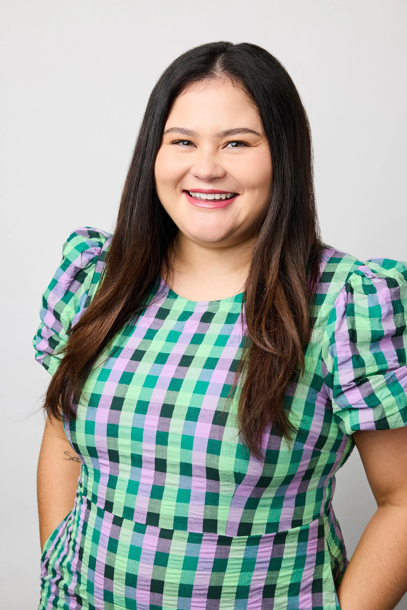 A young woman with long dark hair, smiling, in a green, purple, and black checkered dress, standing against a plain white background.
