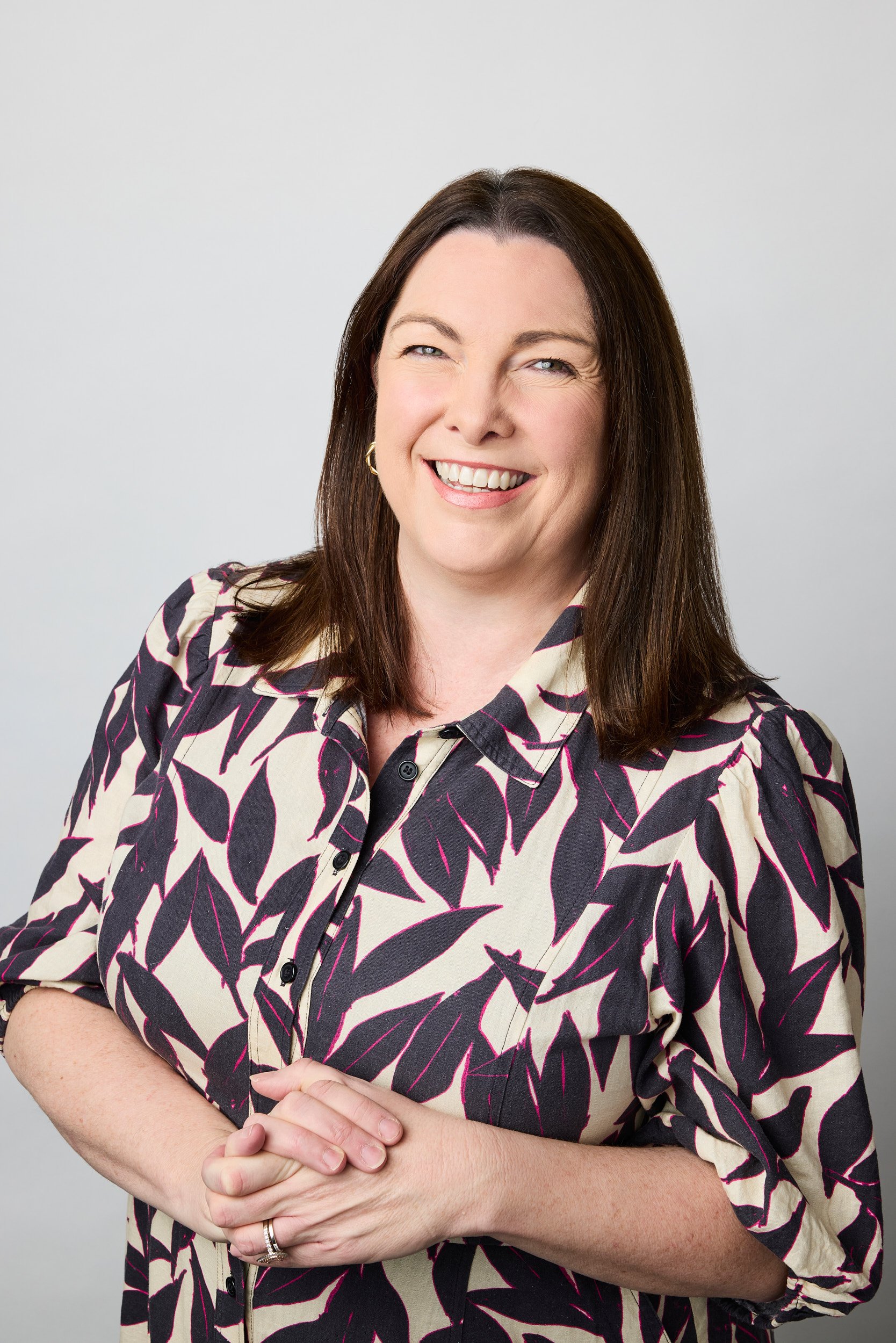 A woman with long brown hair wearing a black, cream, and pink patterned blouse, smiling with her eyes slightly closed, standing against a plain gray background.