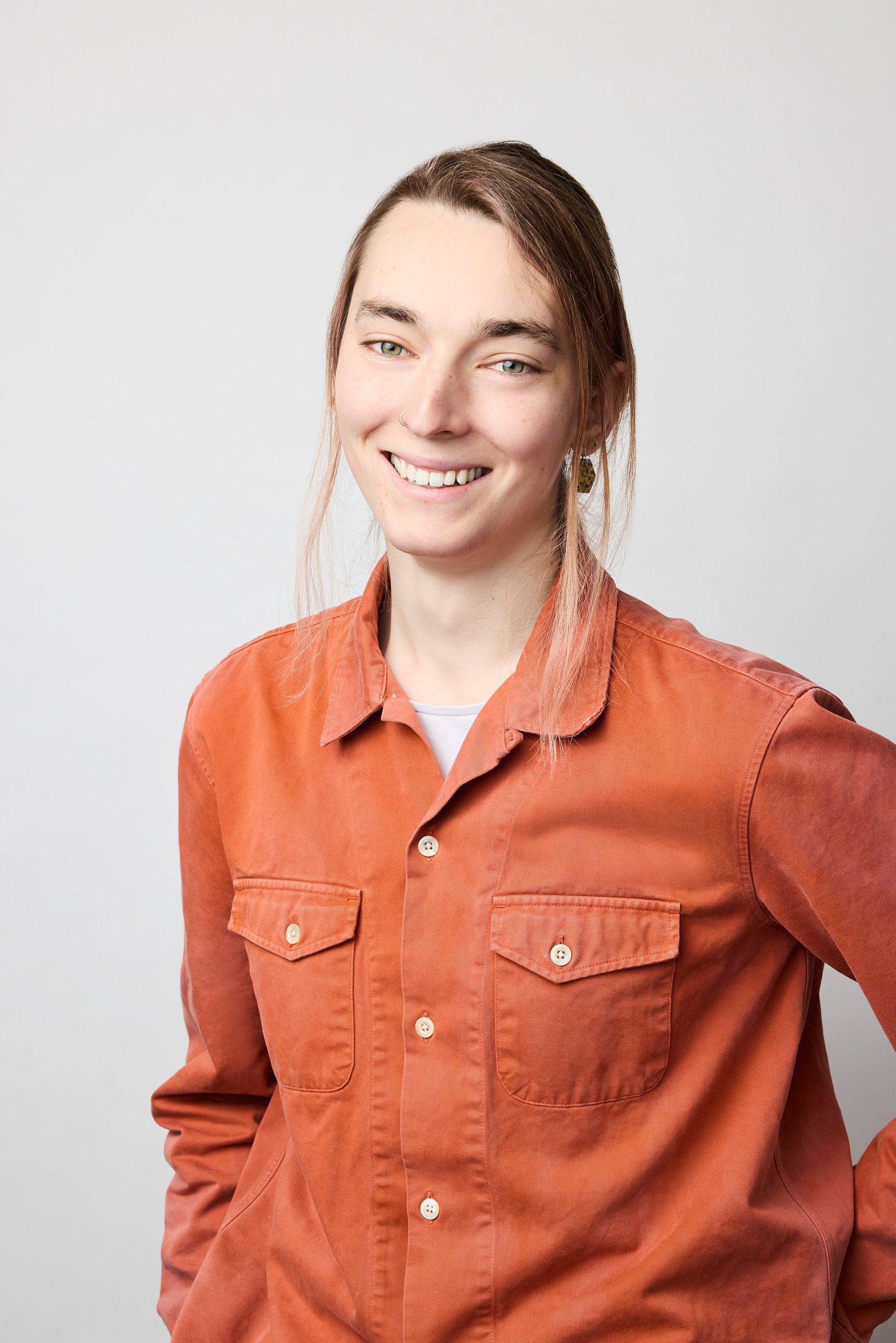 A young woman with long brown hair and light skin, wearing an orange button-up shirt with chest pockets, smiling, and standing against a plain white background.