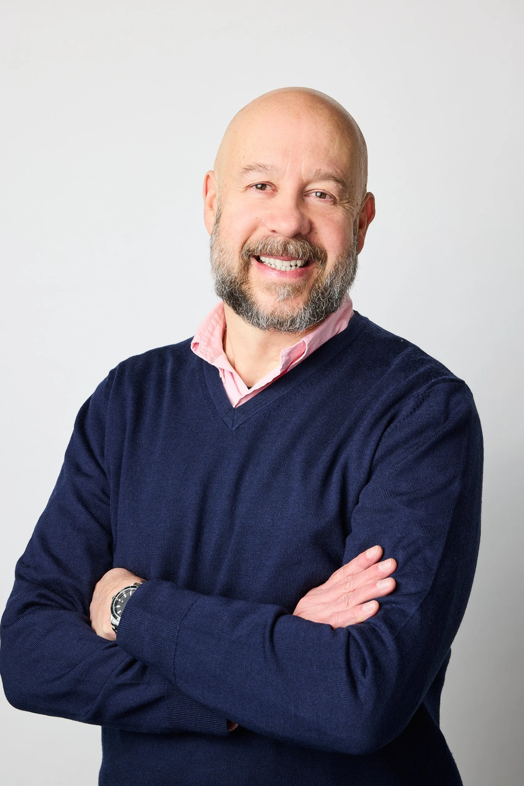 A smiling middle-aged man with a beard, wearing a navy blue sweater over a pink collared shirt, with arms crossed against a plain white background.