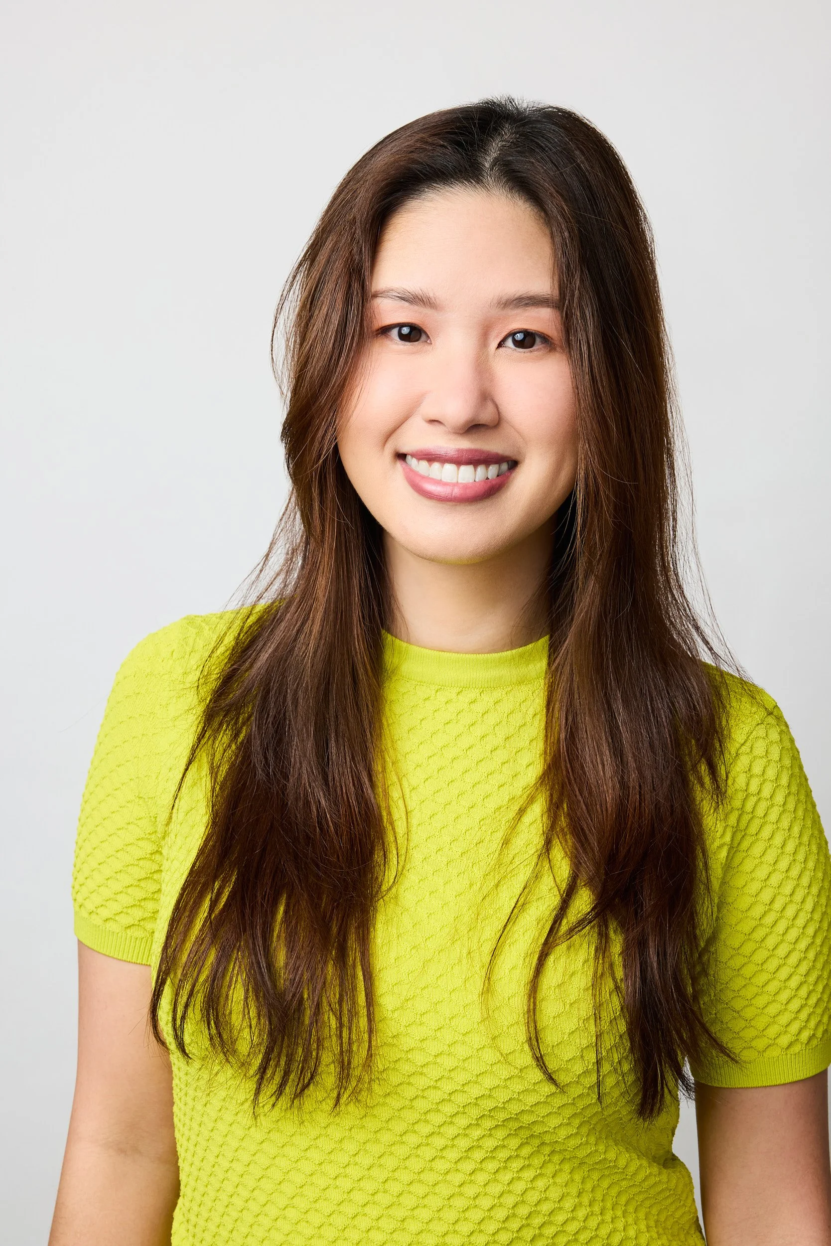 A young woman with long brown hair, smiling, wearing a bright yellow textured short-sleeve shirt against a plain light gray background.
