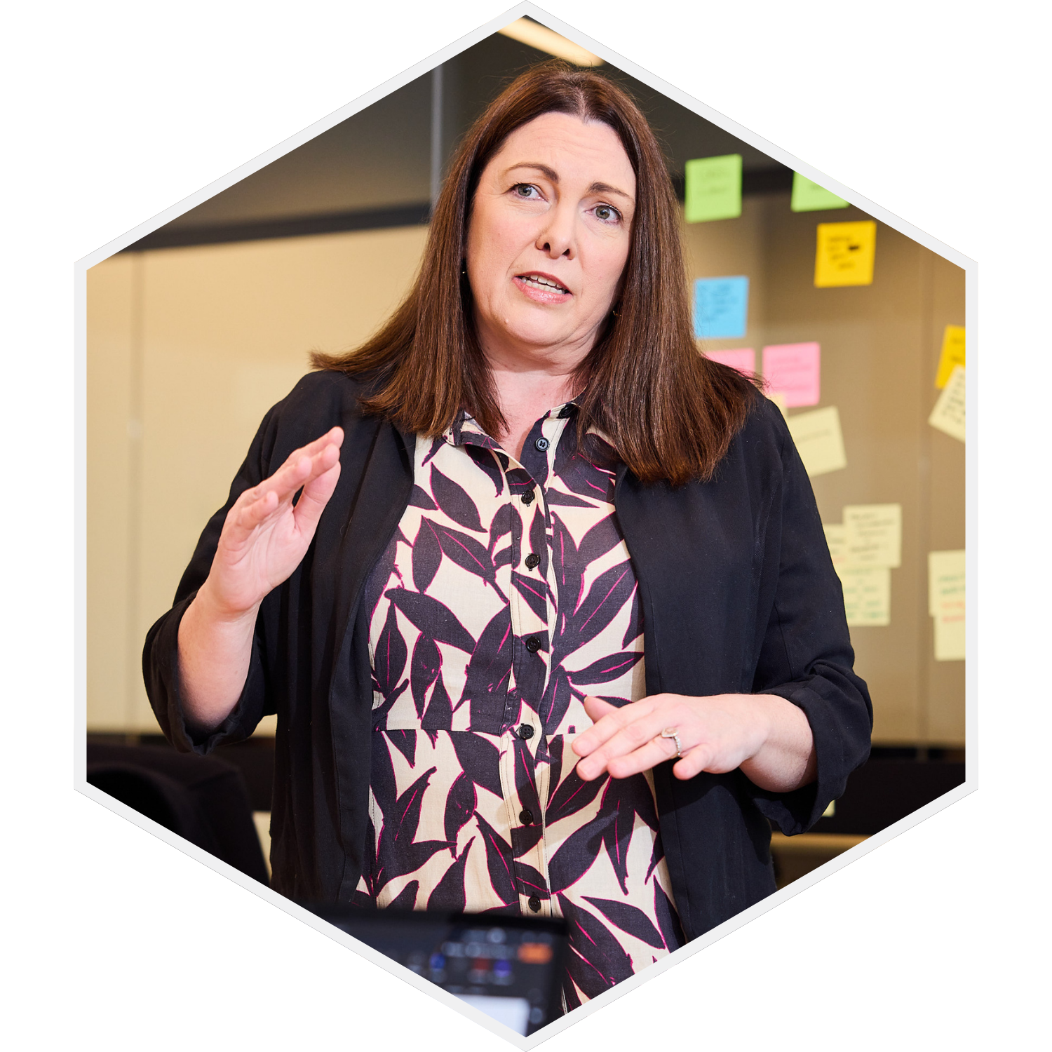 A woman with long brown hair, wearing a patterned blouse and a black jacket, speaking or explaining with her hand raised. Behind her, there are colorful sticky notes on a wall.