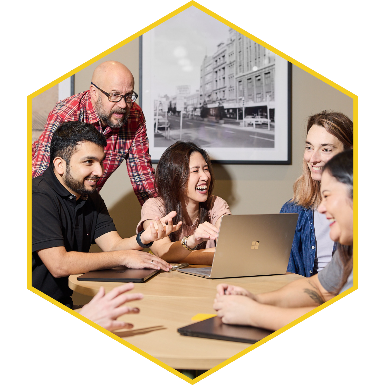 A group of five diverse young people, three women and two men, are gathered around a laptop on a wooden table, laughing and engaging in conversation. An older man with glasses stands behind them, leaning over to join the discussion. The background features a framed black-and-white cityscape photograph on the wall.