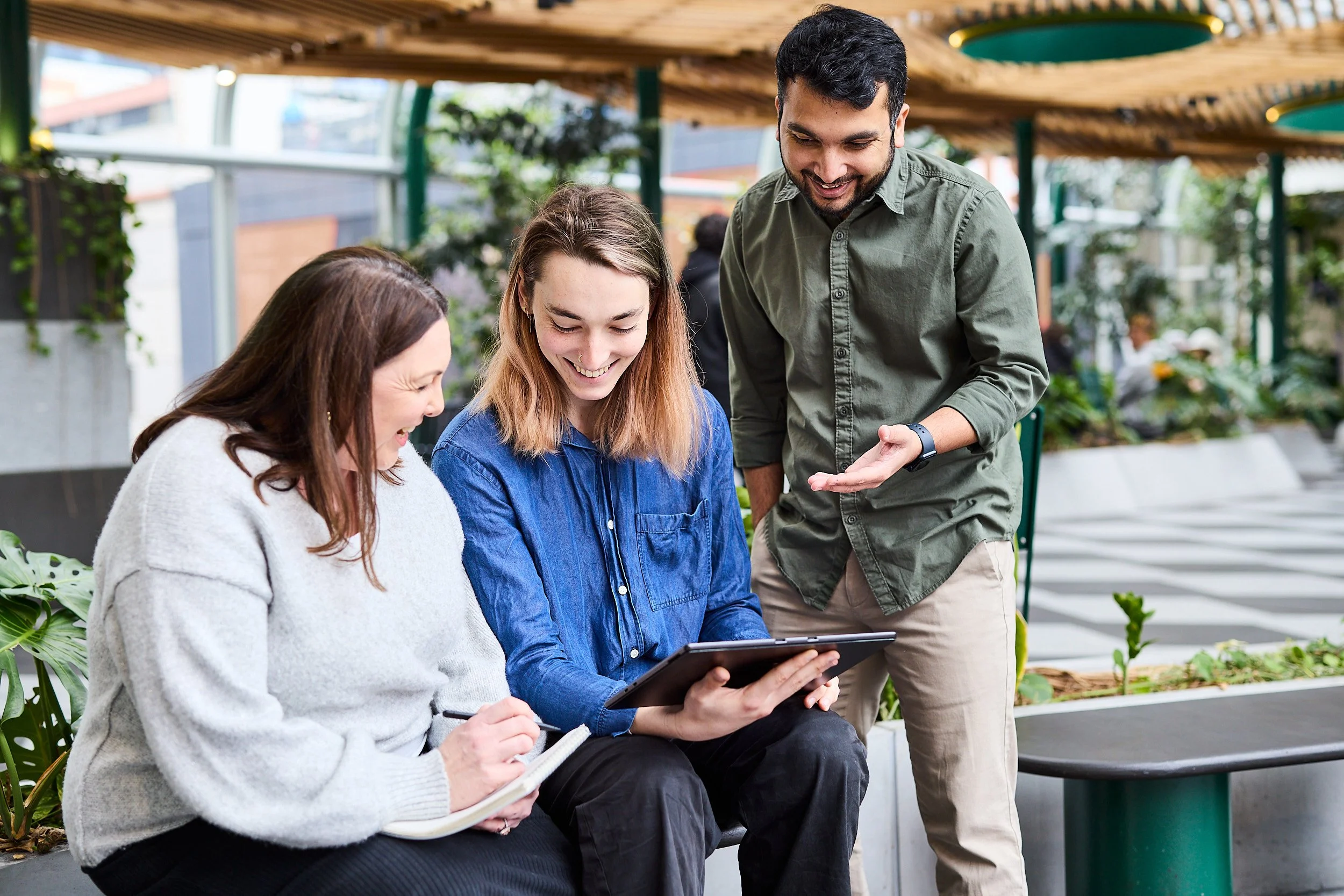 Three people, two women and one man, are gathered outside in a garden center or greenhouse. The women are seated, one holding a notebook and pen, the other looking at a tablet, while the man stands next to them, smiling and gesturing towards the tablet.