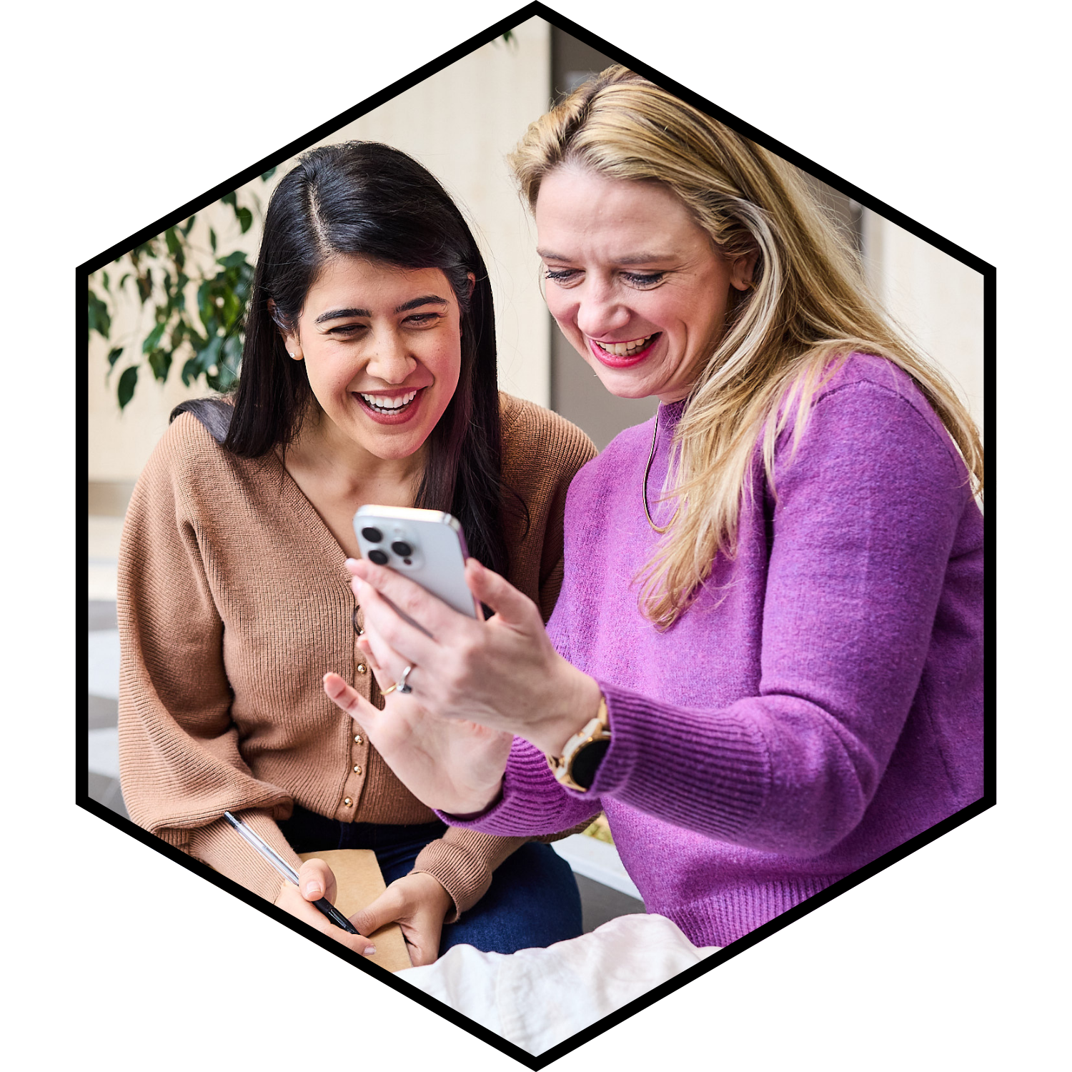 Two women smiling and looking at a smartphone together, one holding a pen and notepad.