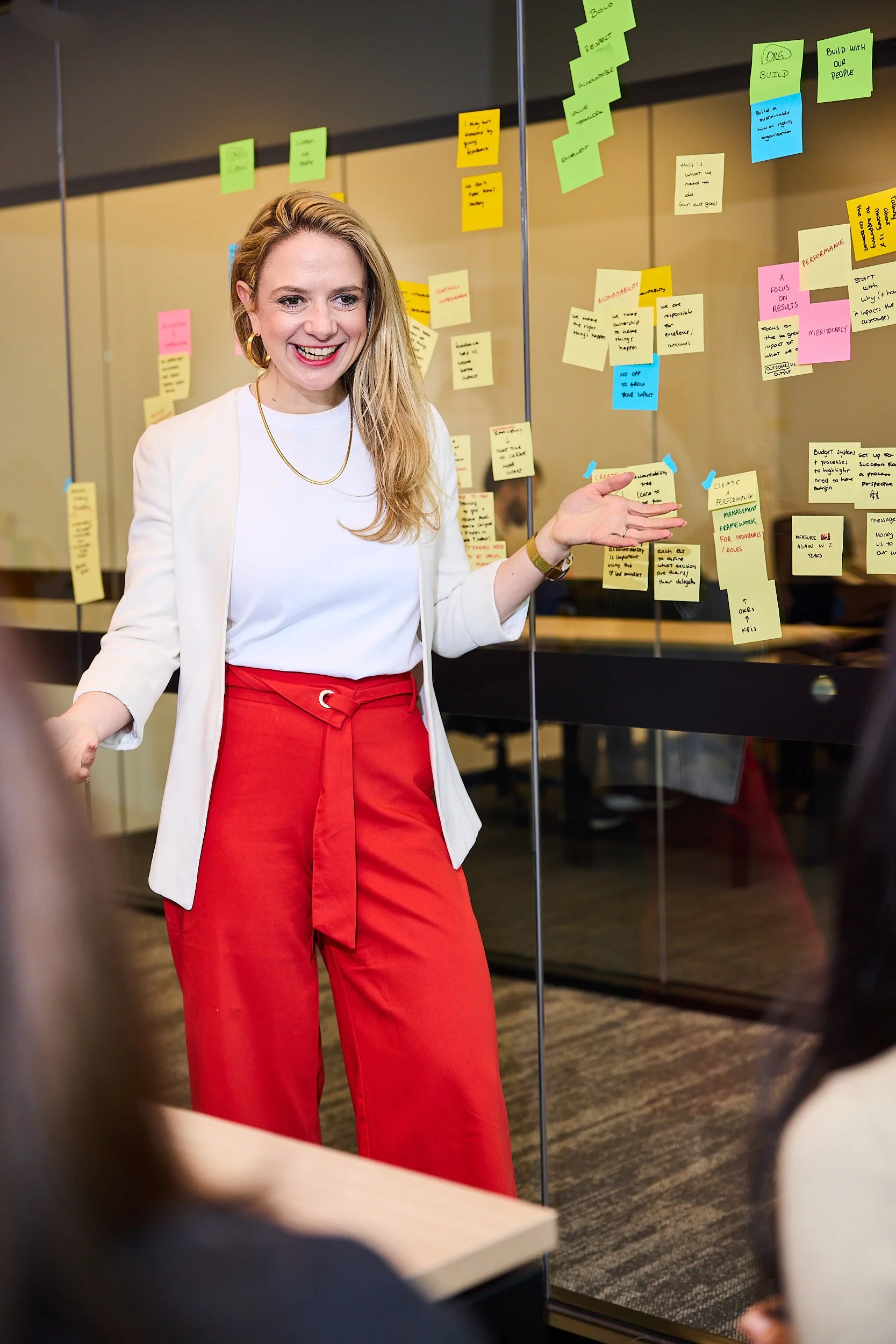 A woman smiling and gesturing towards a glass wall covered with colorful sticky notes, in a professional meeting or workshop setting.
