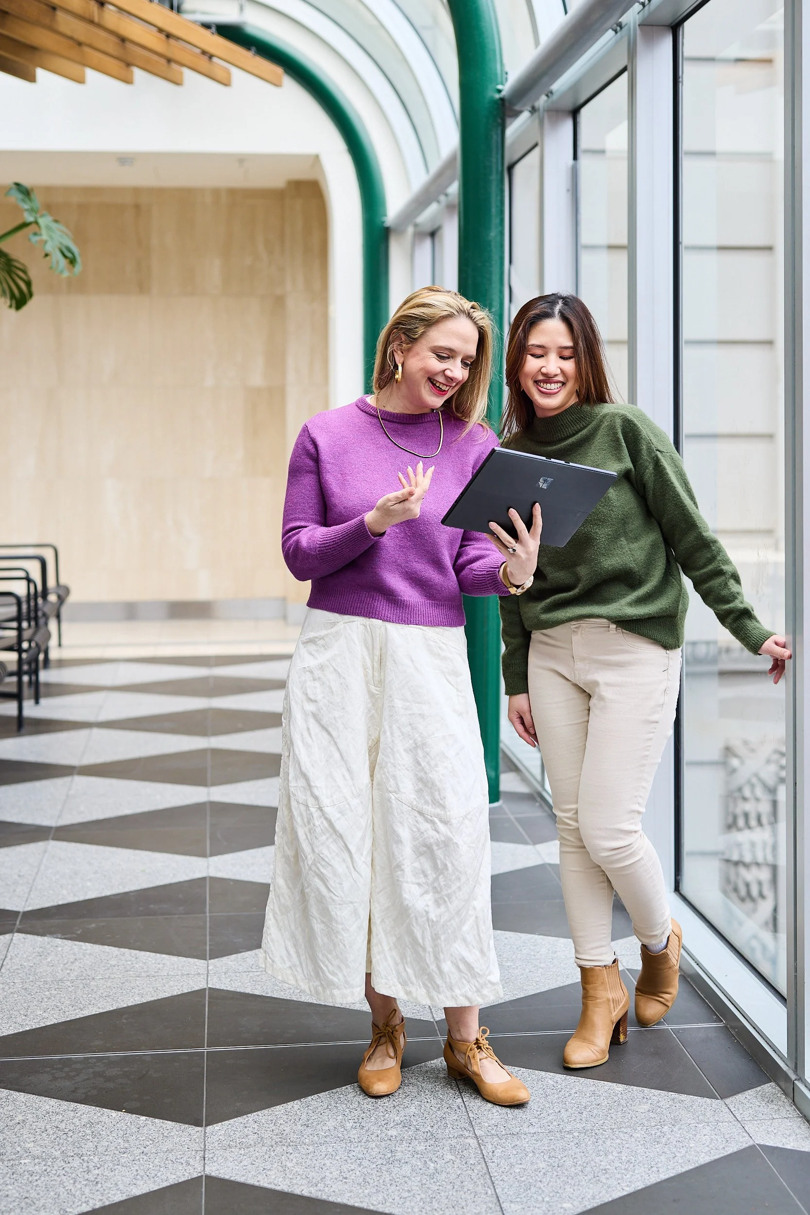Two women smiling and looking at a tablet inside a modern building with glass walls and black-and-white checkered floor.