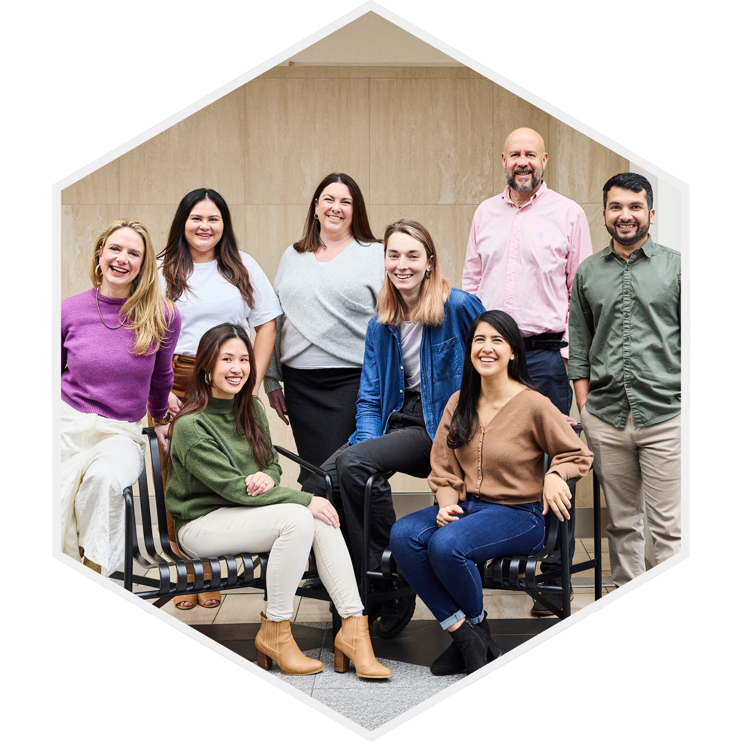 A diverse group of nine people, including women and men, posing together indoors with a beige wall in the background. They are smiling and appear to be in a casual or professional setting.